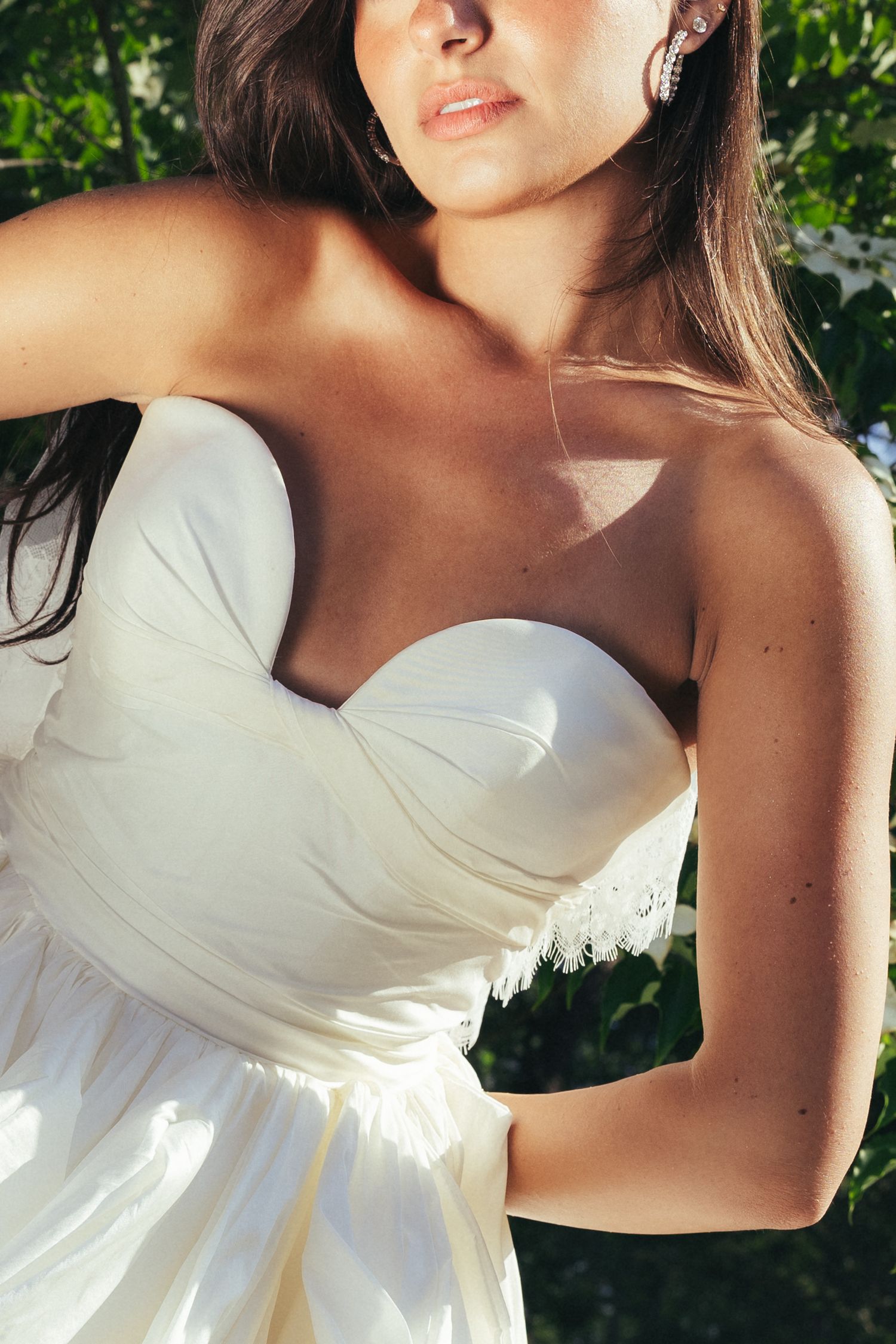Close-up of bride in strapless white wedding dress with lace detail posing outdoors in sunlight.