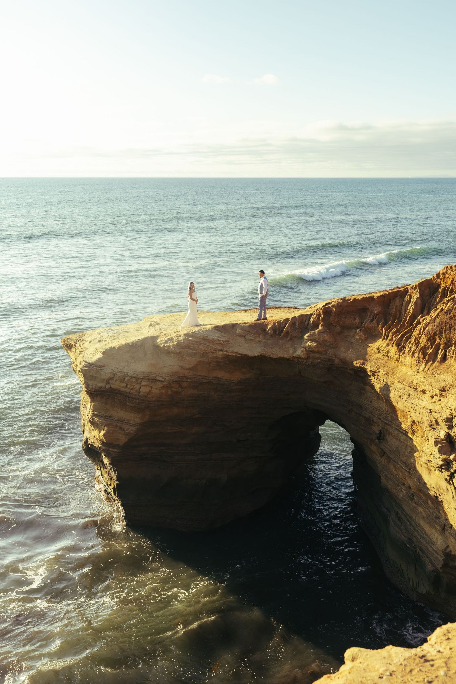 Couple standing on a rocky coastal cliff arch overlooking the ocean at sunset in a romantic seaside setting.