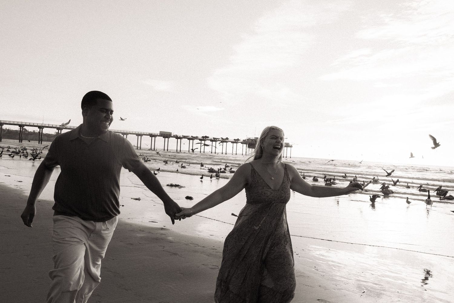 Couple holding hands walking on beach near pier, black and white romantic photo with seagulls flying.