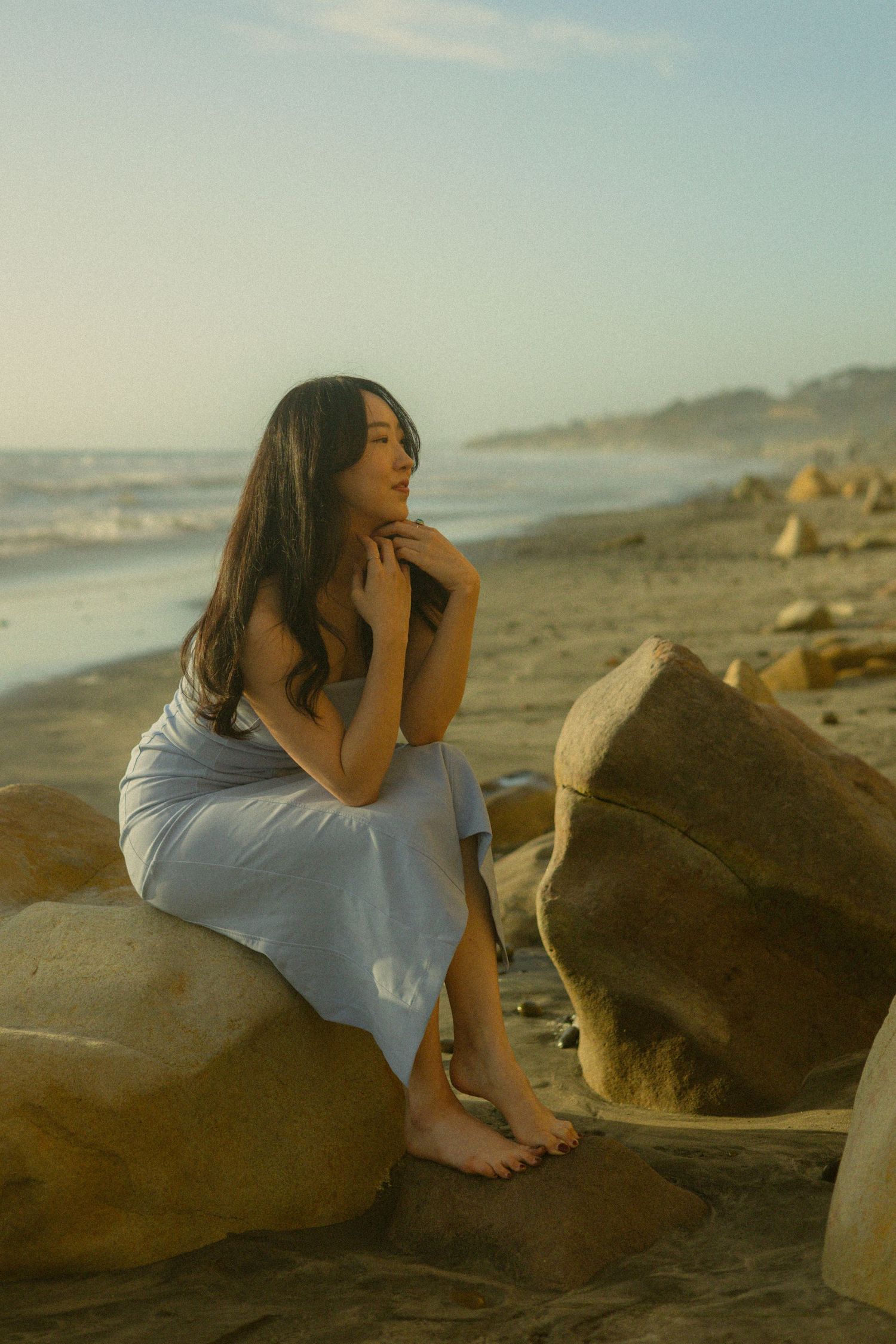 A pensive woman in a white dress sits on a large rock at sunset on a misty beach.