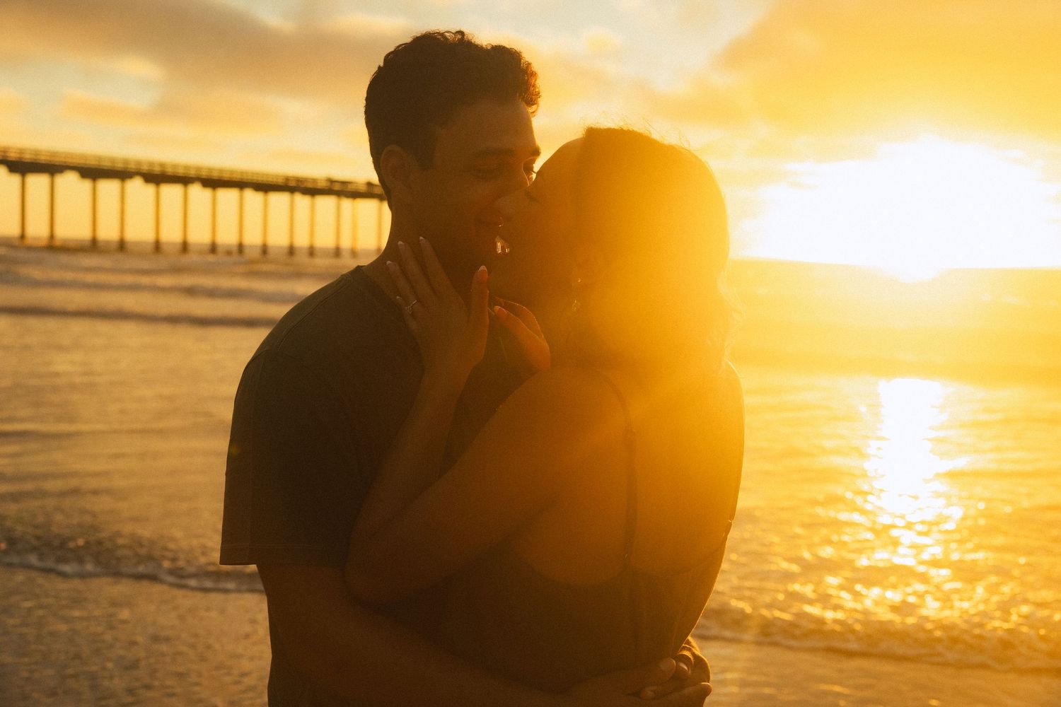 A silhouetted couple embraces on a beach at sunset with a pier in the background.