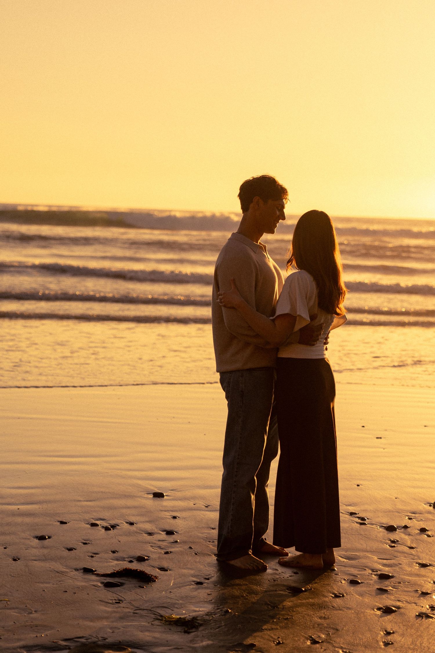 A couple shares an intimate moment on a beach at sunset with golden light reflecting off the ocean waves.
