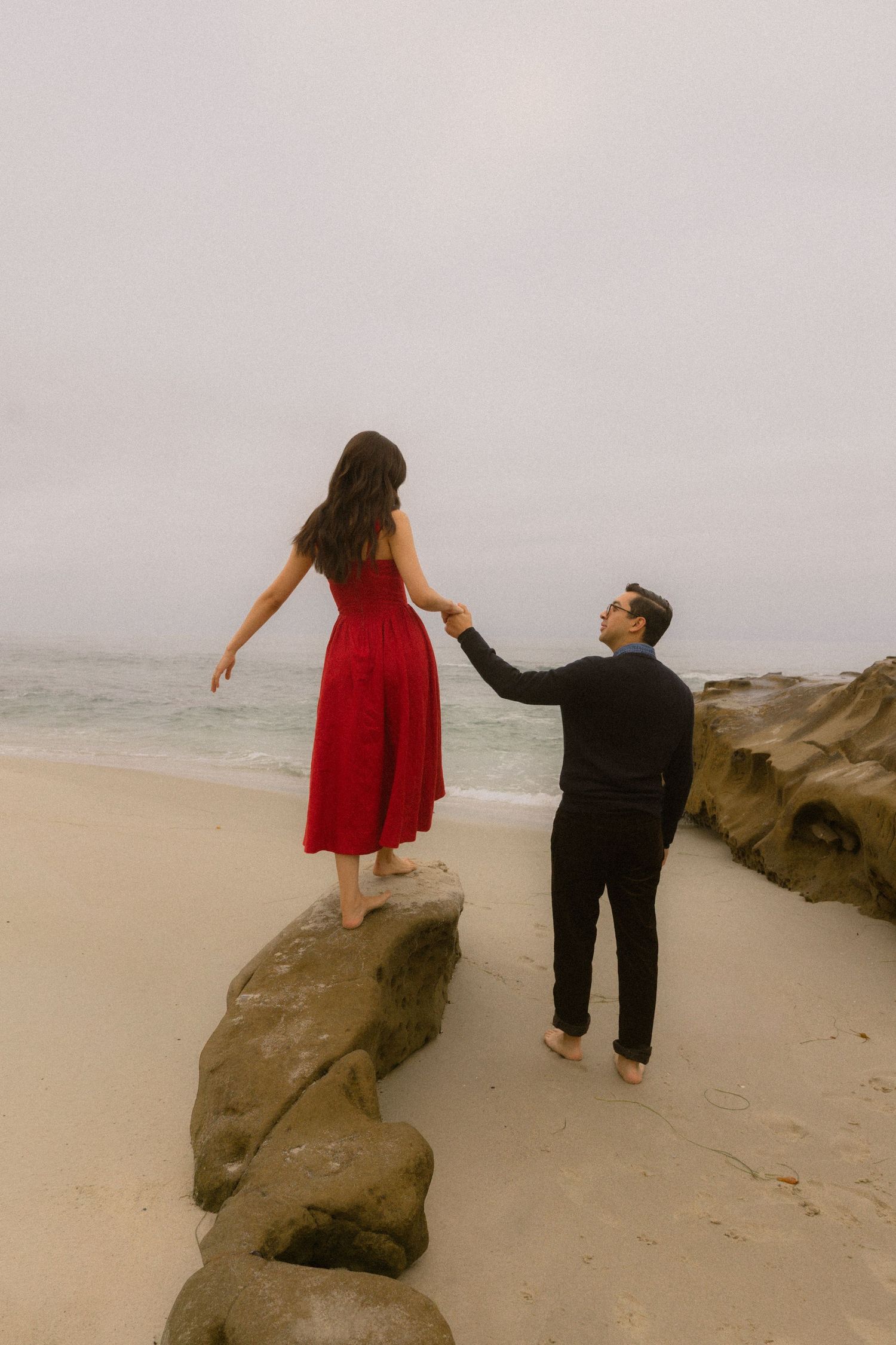 Two people in casual attire stand on large rocks at a misty beach, creating playful poses against the foggy backdrop.
