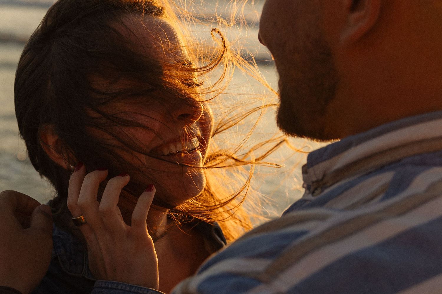 Couple sharing a romantic moment at golden hour, woman smiling with wind-blown hair at the beach.