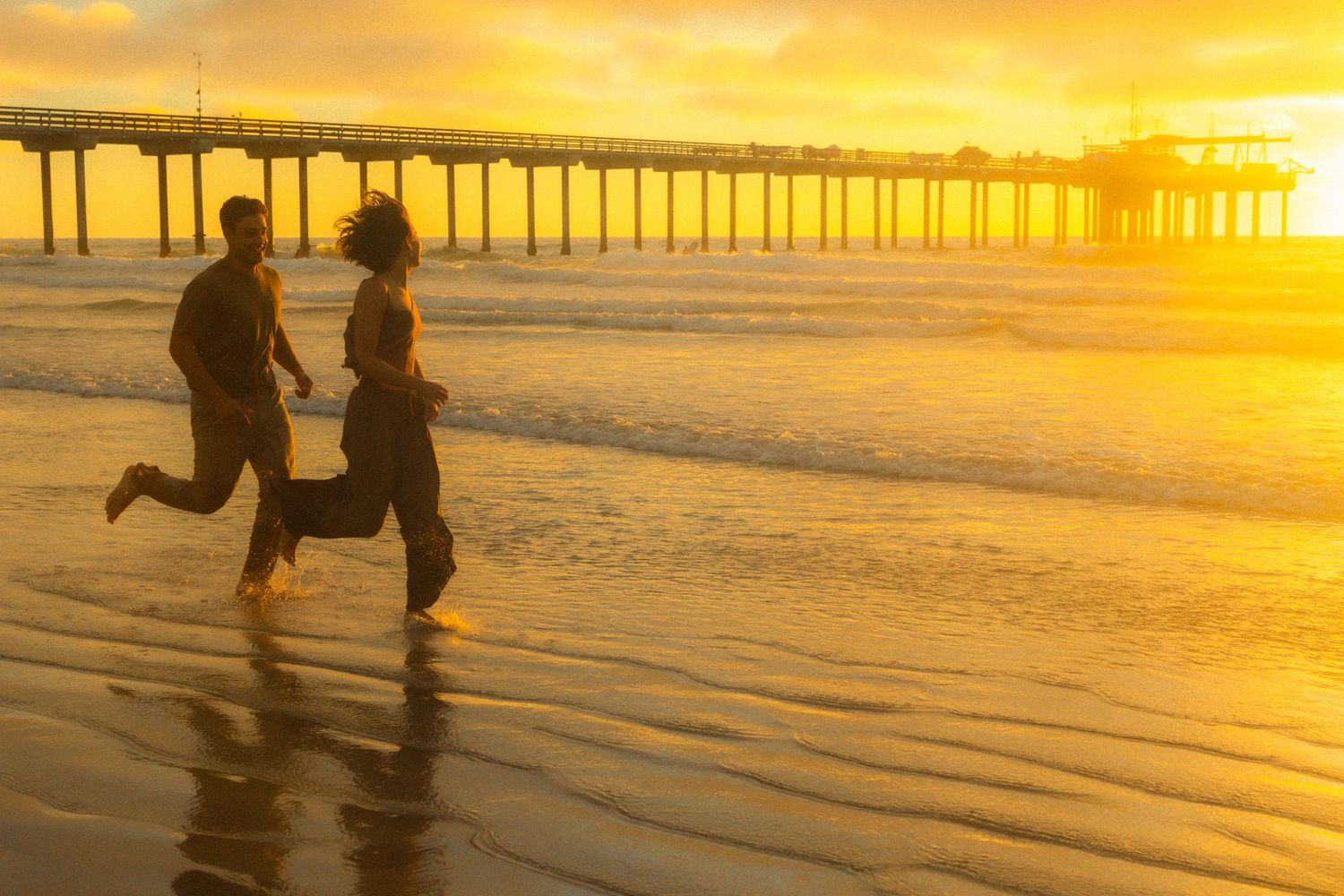 Silhouettes of people walking along a beach at sunset with a wooden pier in the background.