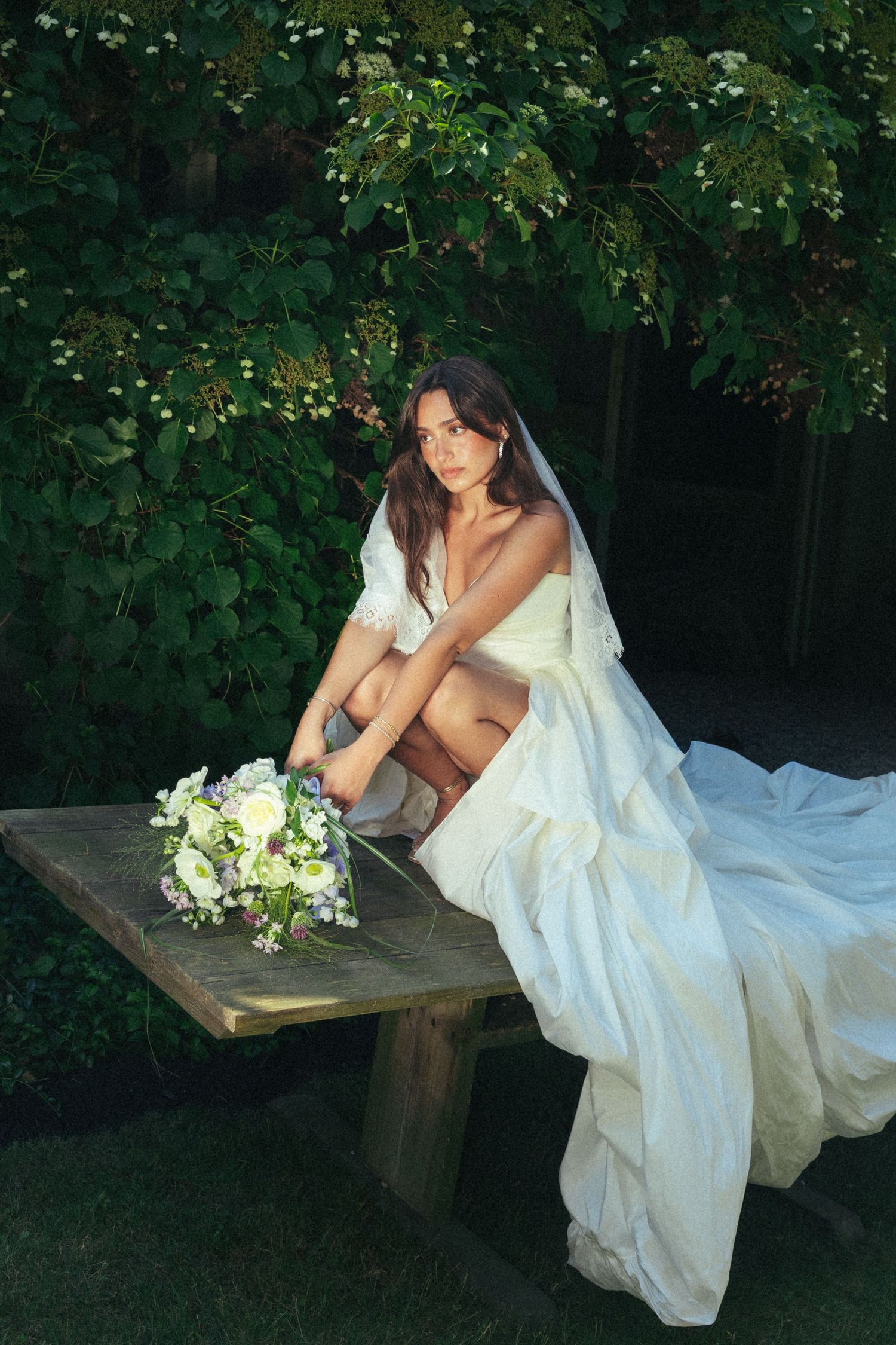 Elegant bride in a white off-shoulder gown sitting on a wooden bench with a floral bouquet in a lush garden.