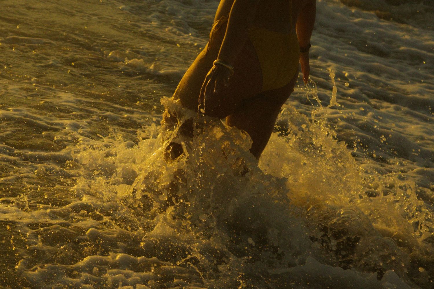 Horse hooves splash through shallow water at sunset creating golden droplets in backlit sunlight.