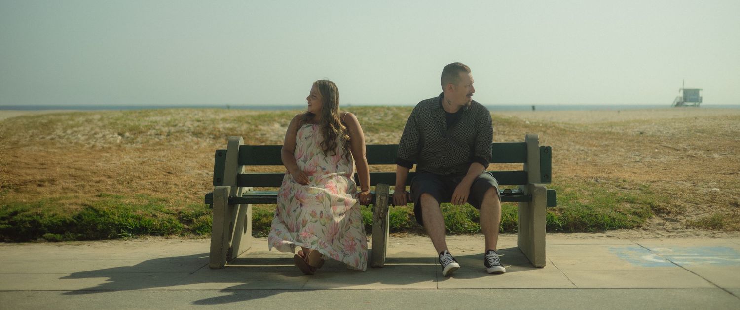 Two people sit apart on a wooden bench overlooking a coastal landscape on an overcast day.