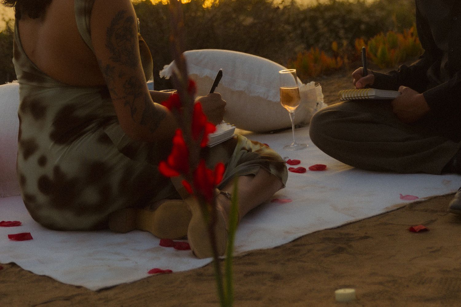 A romantic picnic setup at sunset with wine glasses and a white blanket spread on the ground.