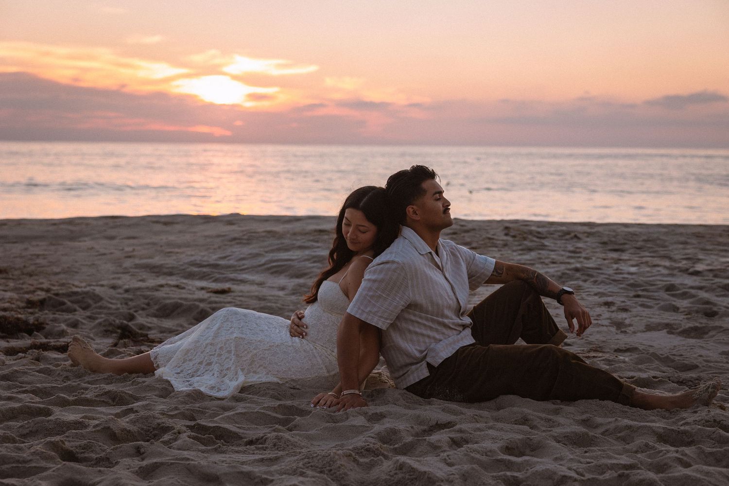 A romantic couple in wedding attire embraces on a sandy beach at sunset with orange and pink sky in the background.