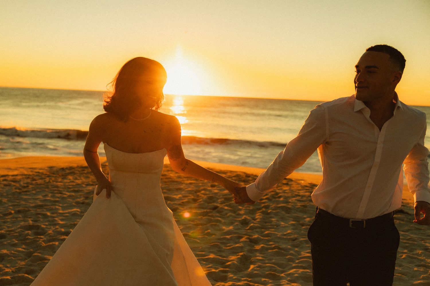 Couple walking on beach at sunset holding hands with golden sunlight creating silhouettes on sandy shore.