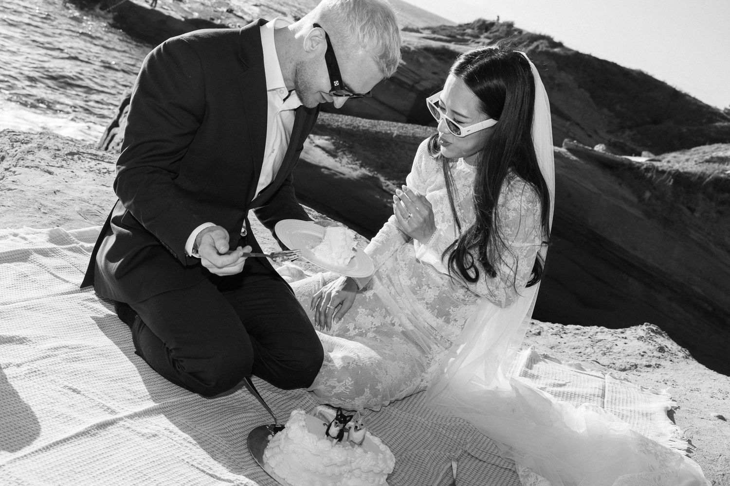 Black and white photo of a bride and groom exchanging vows on a rocky beach, bride in lace gown.