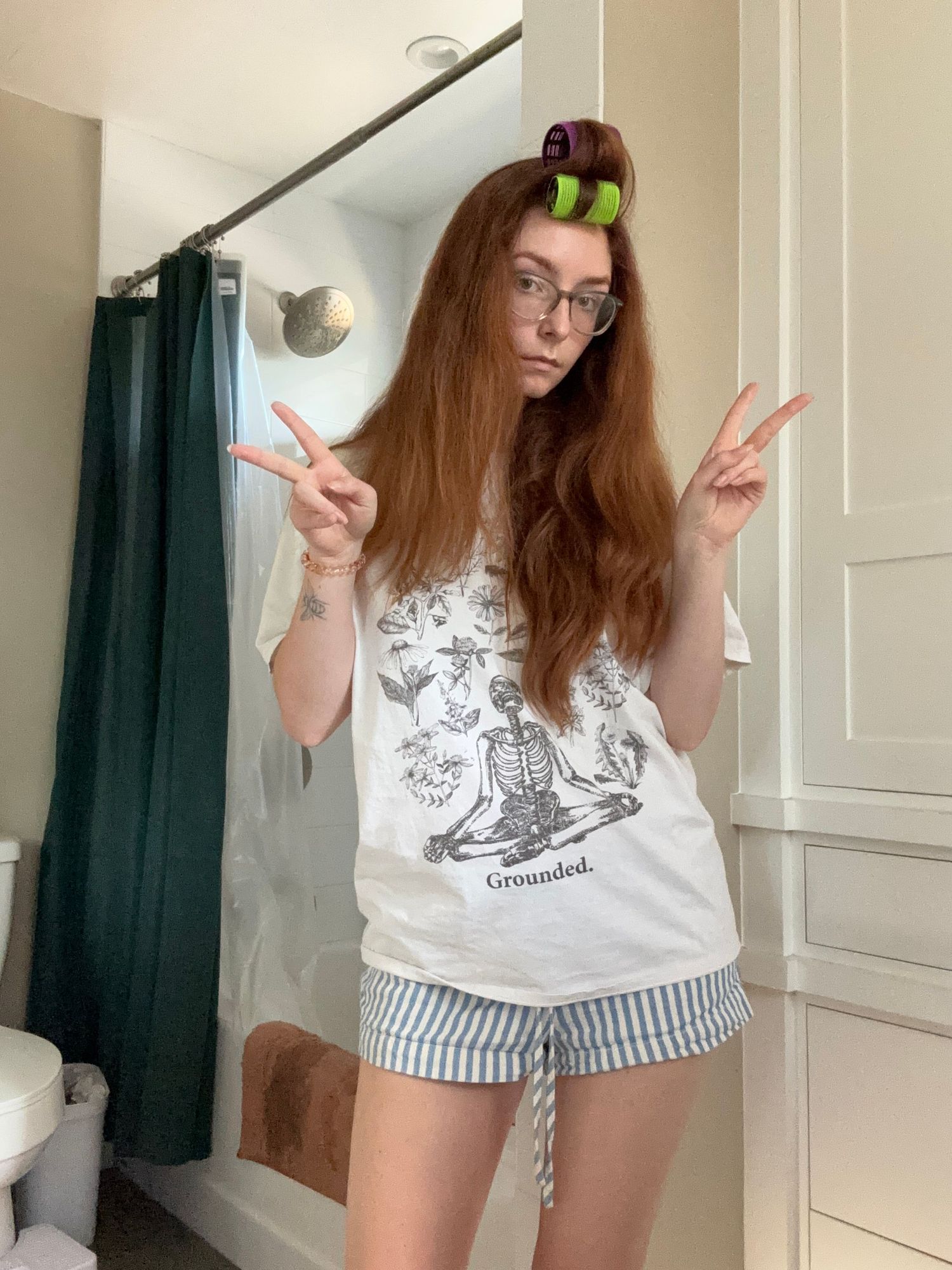 Young woman with long red hair and hair rollers making peace signs, wearing a skeleton graphic tee and striped shorts in a bathroom.