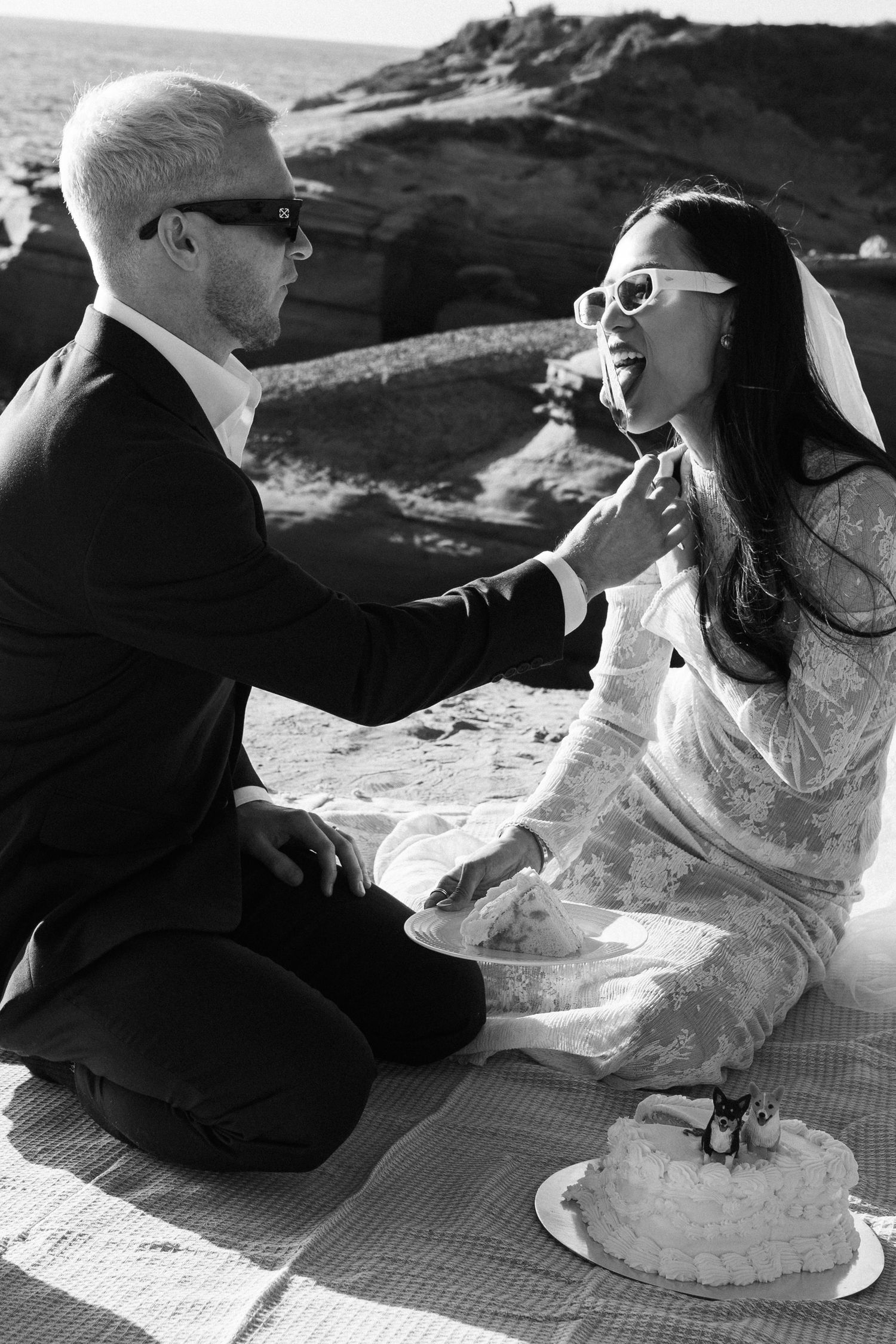 Black and white photo of bride and groom sharing wedding cake on beach blanket, wearing sunglasses.