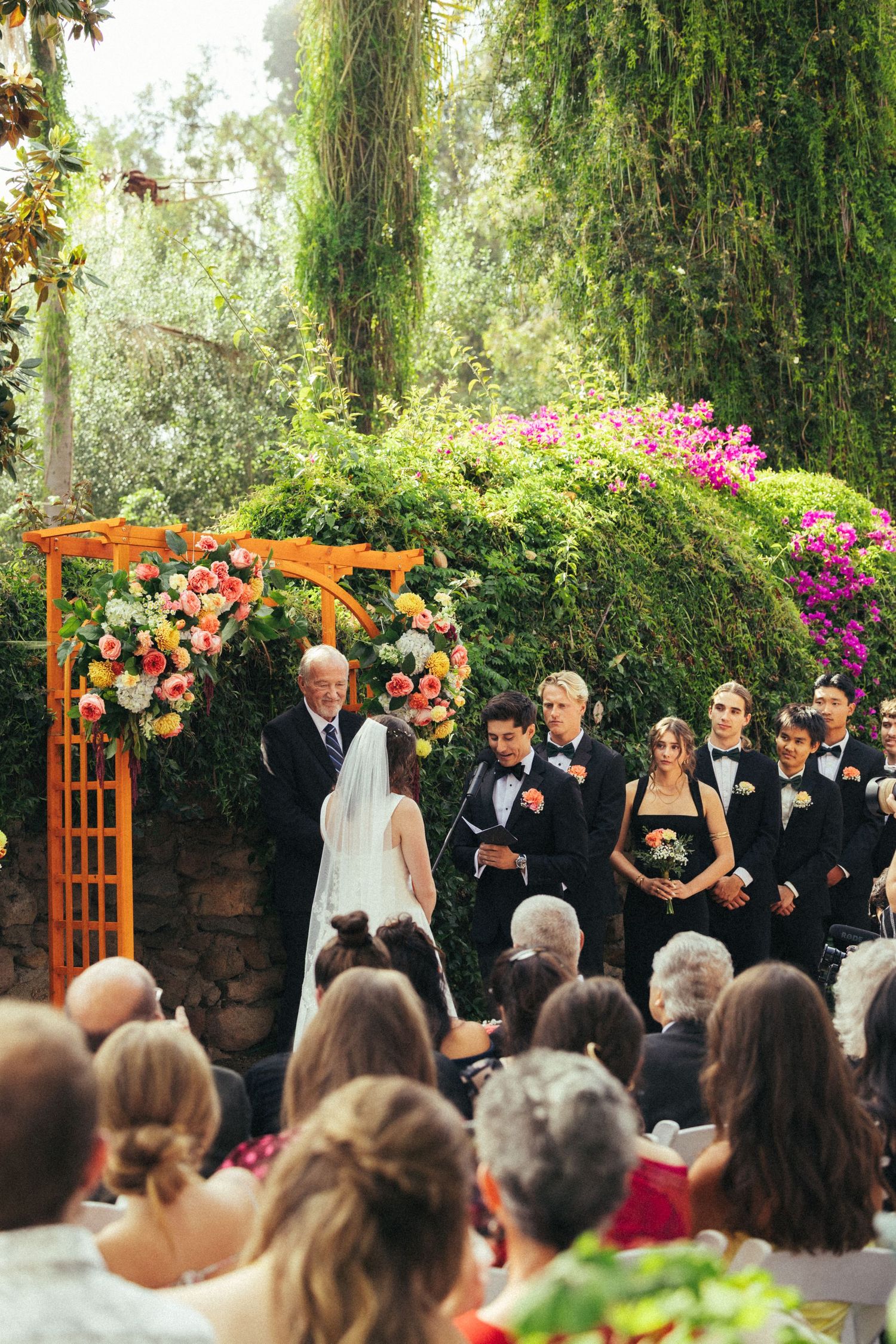 Outdoor wedding ceremony with couple at floral arch, wedding party in tuxedos, lush green ivy wall and colorful flowers.