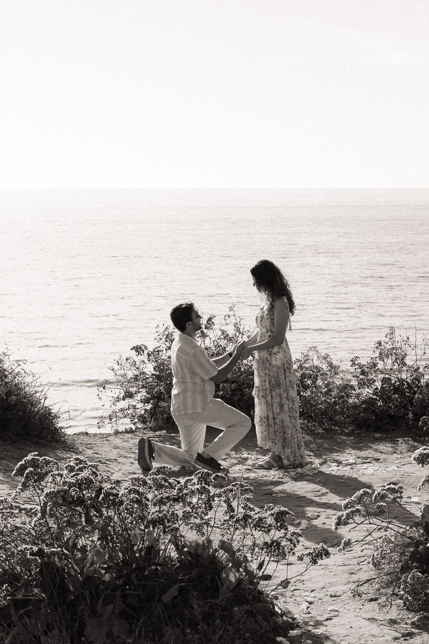 Black and white photo of a man on one knee proposing to a woman on a cliff overlooking the ocean.