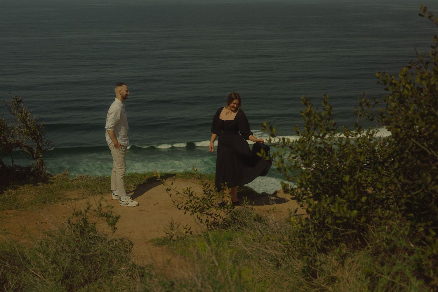 Couple standing on coastal cliff overlooking ocean waves below.