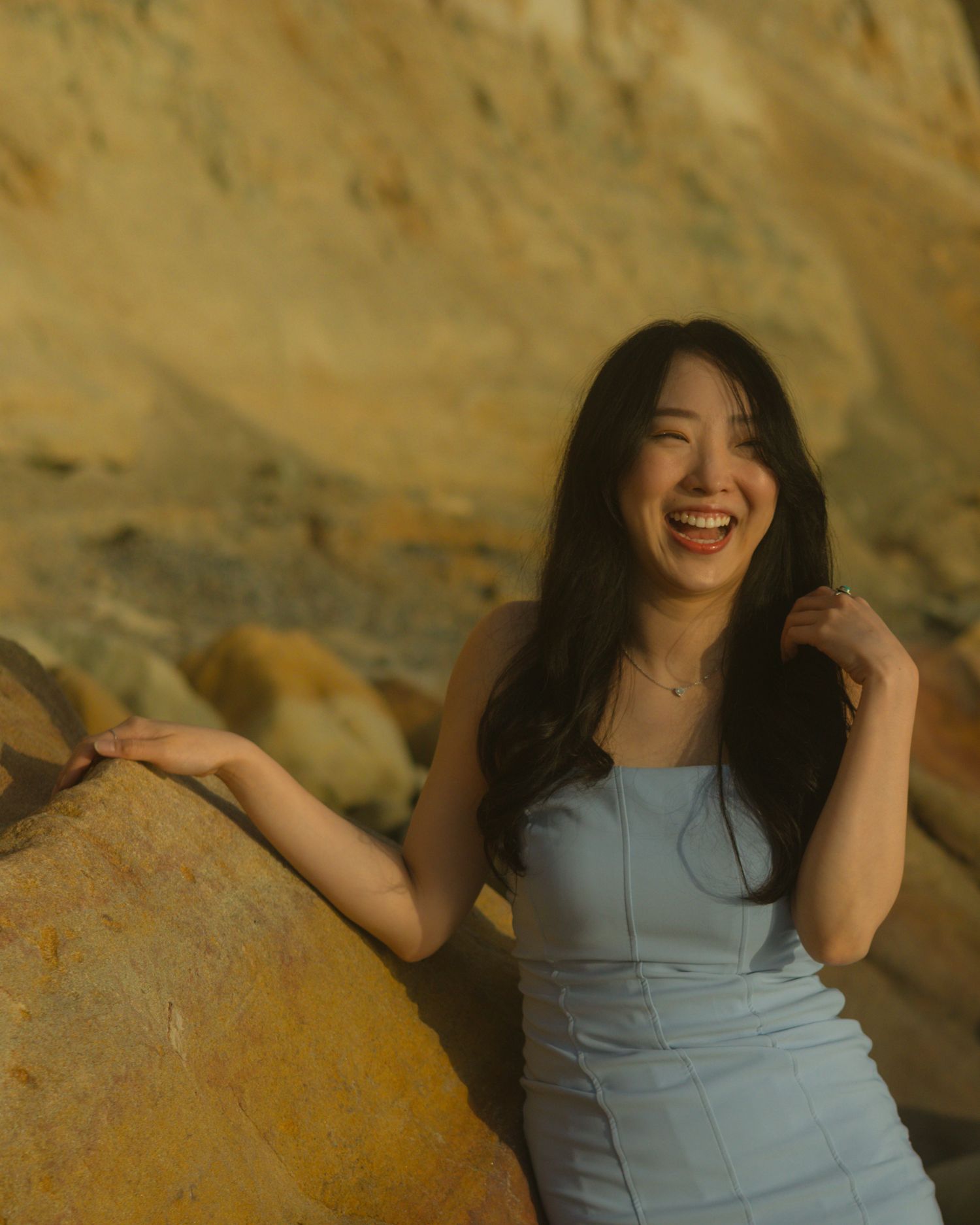 Woman in light blue dress laughs joyfully while posing against golden rocky backdrop during sunset.