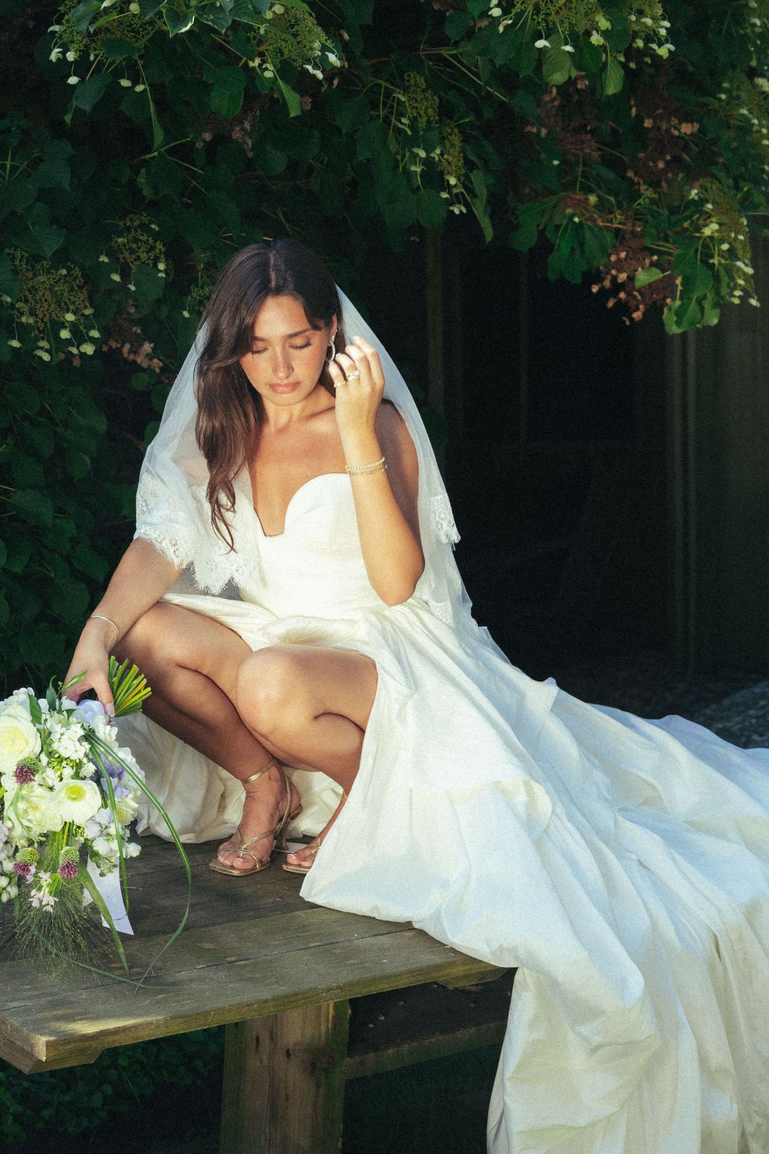 Elegant bride in white dress and veil seated on bench with floral bouquet outdoors.
