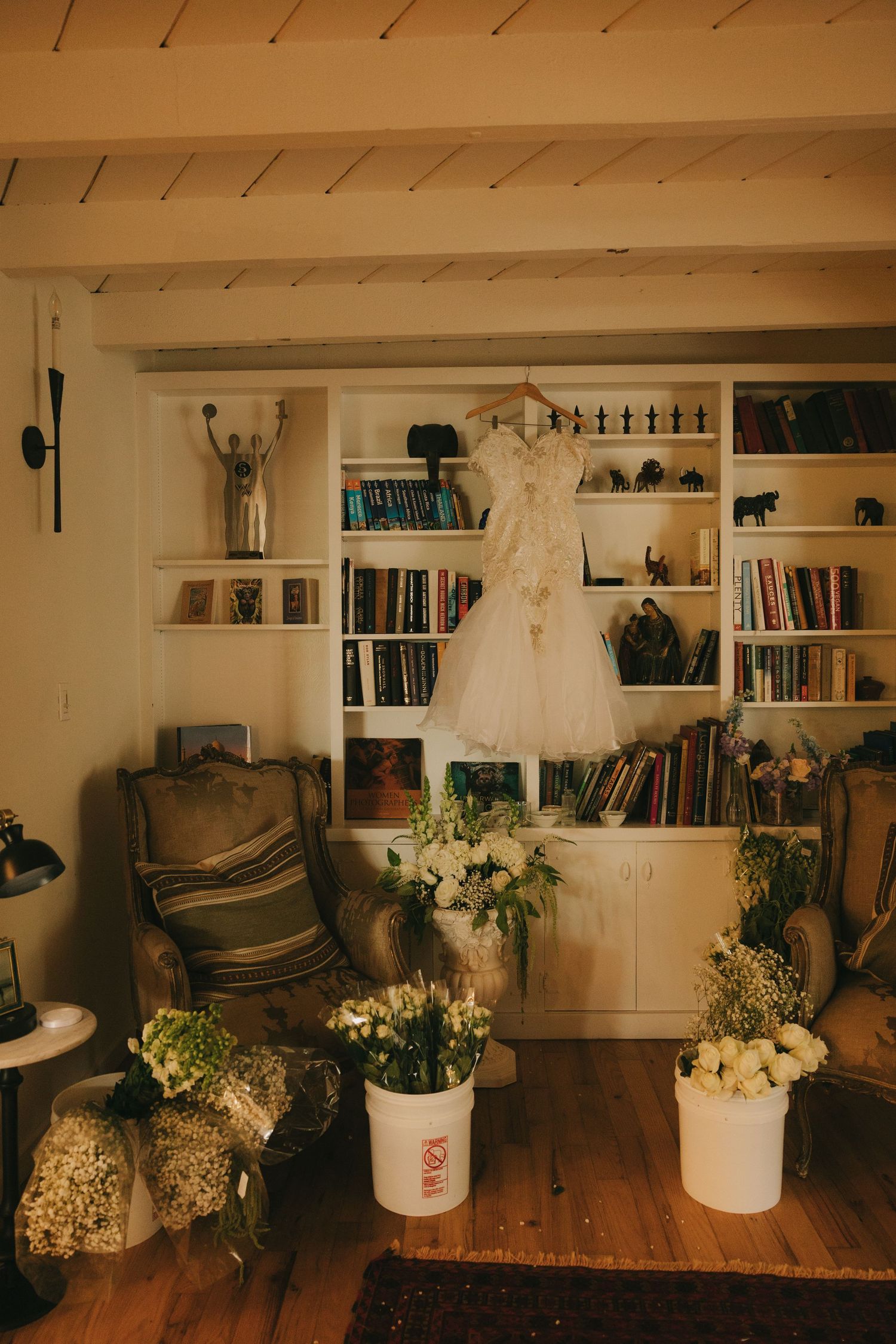 Wedding dress hanging on white bookshelf surrounded by fresh flower arrangements in rustic cozy room with hardwood floors.