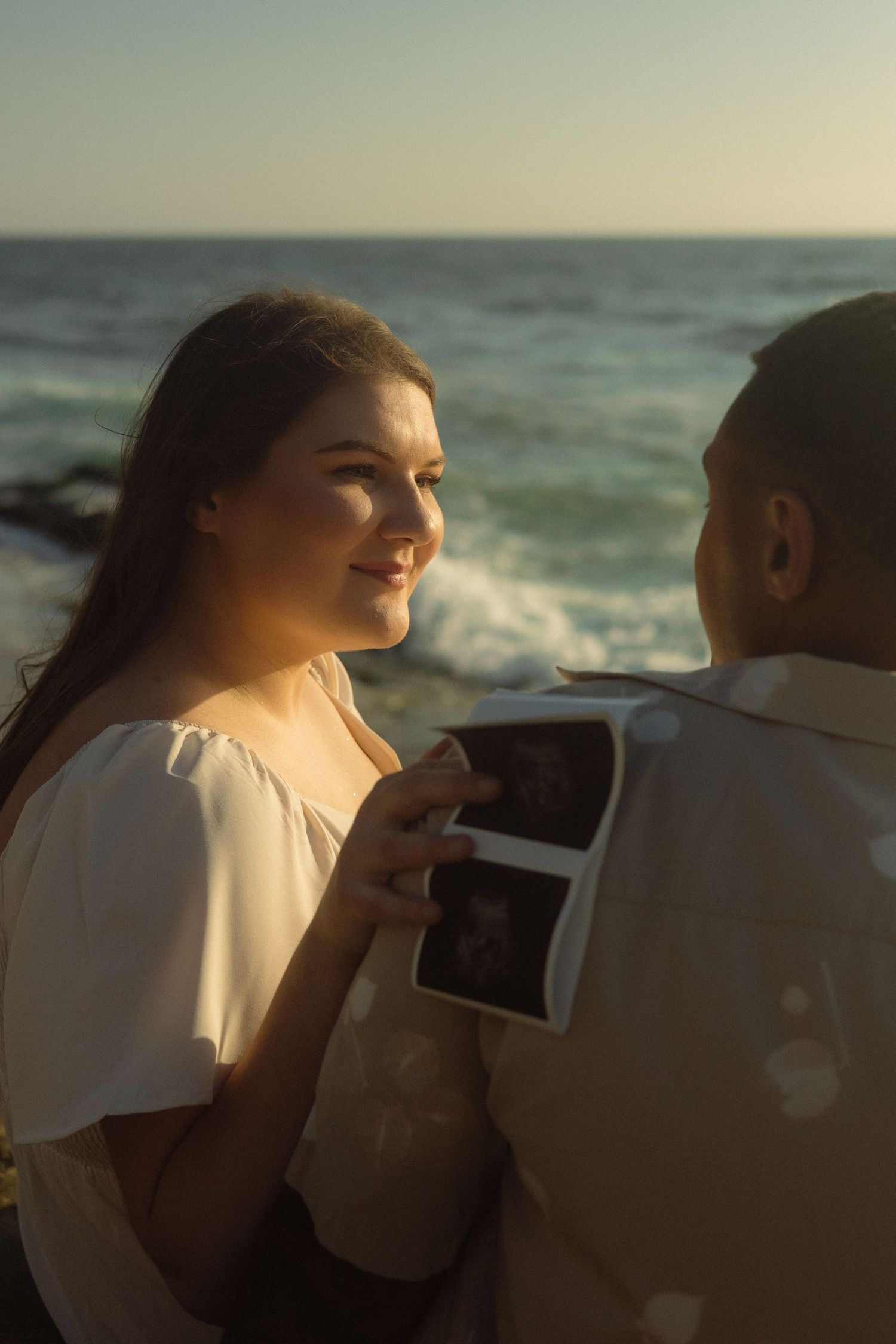 Couple at sunset sharing a special moment by the ocean as they look at what appears to be a sonogram photo.