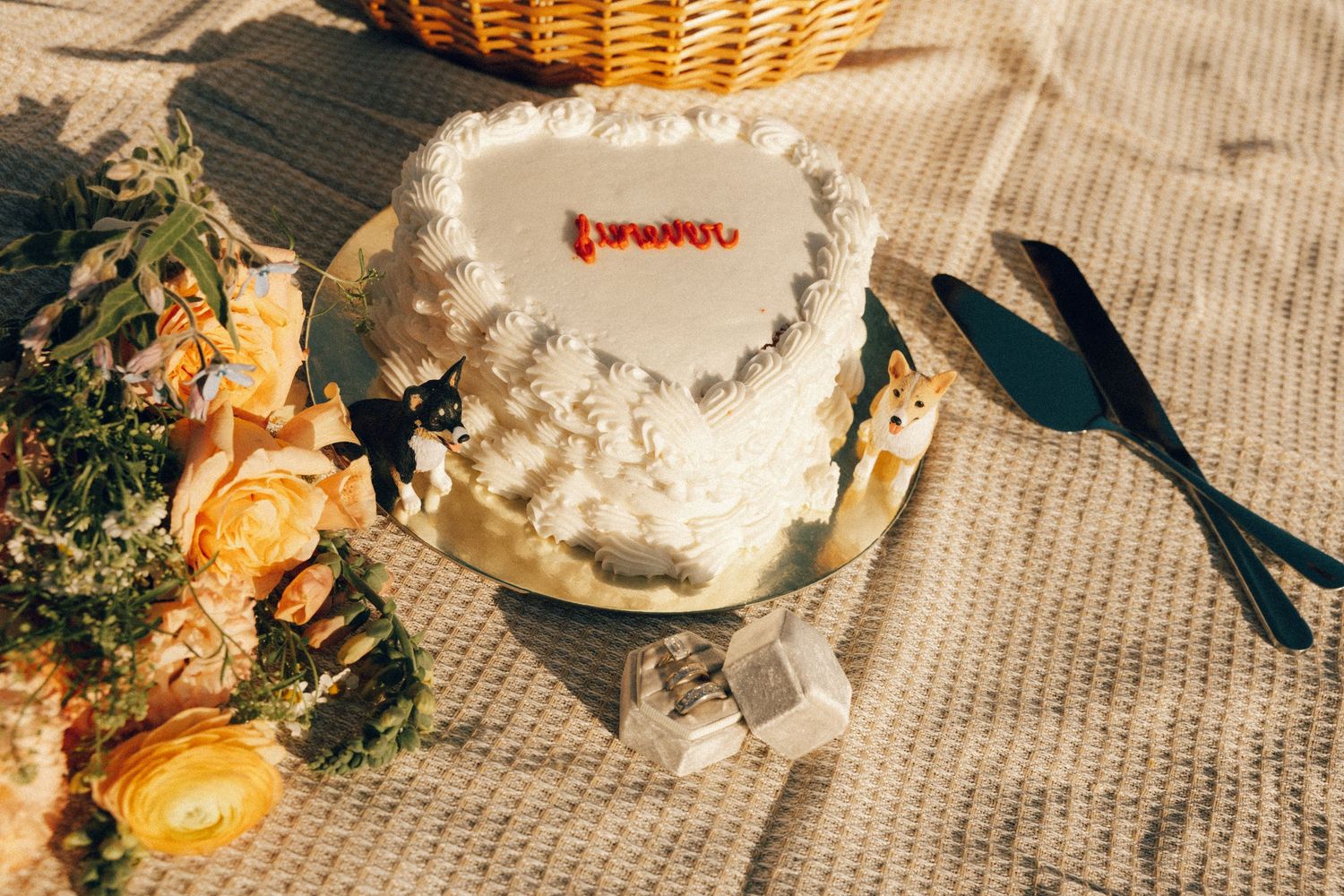 White heart-shaped wedding cake with red lettering on a gold stand, surrounded by yellow flowers and cake servers.