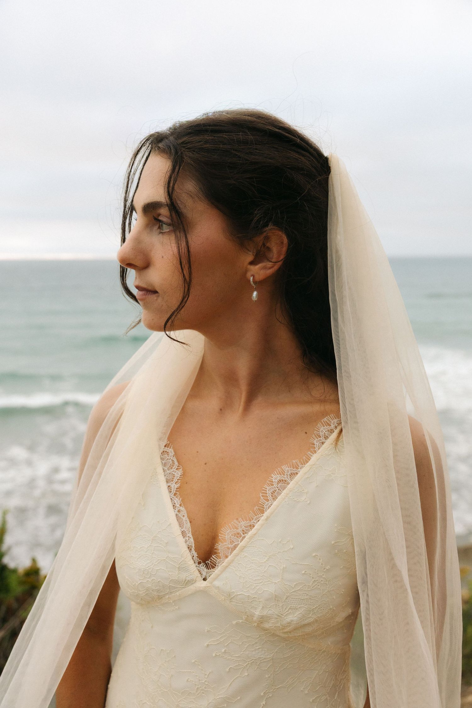 Bride in lace wedding dress and veil gazing oceanside on overcast day.
