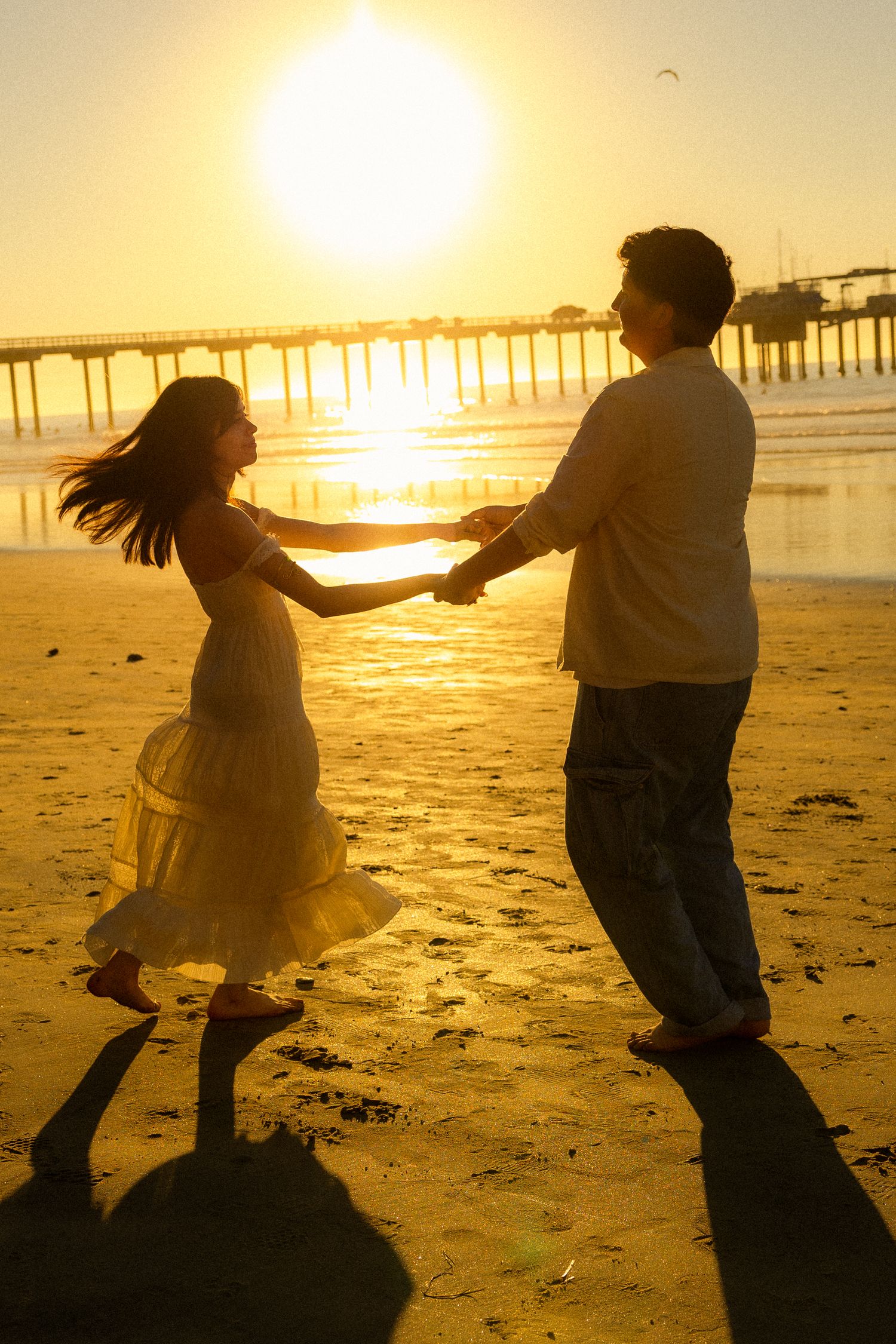 Silhouettes of two people dancing joyfully on a beach at sunset with a pier in the background.