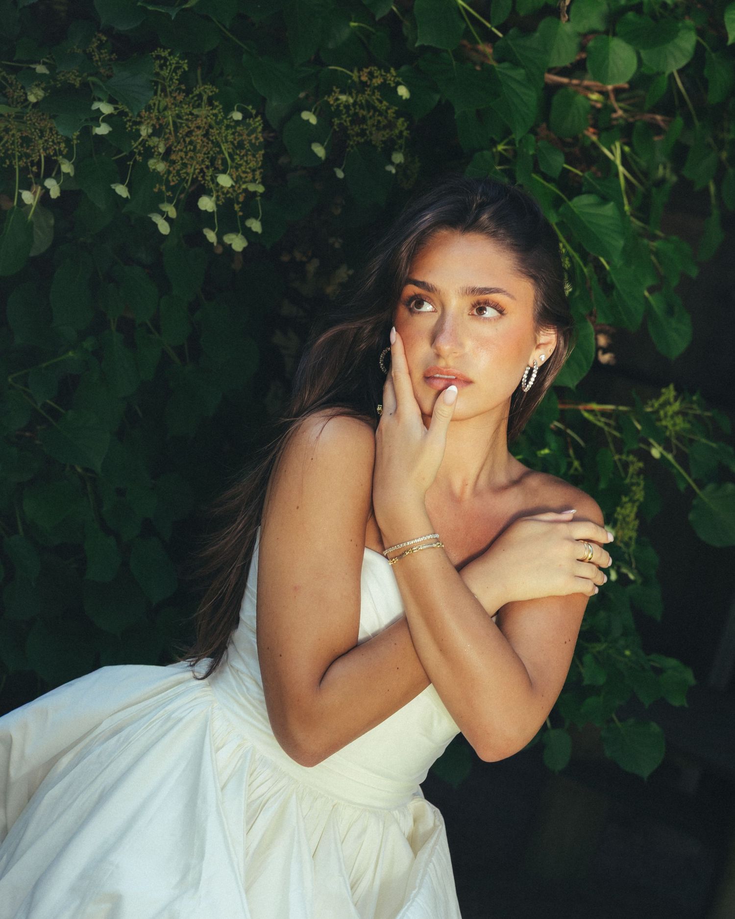 Brunette bride in strapless white gown poses thoughtfully among green leafy foliage outdoors.