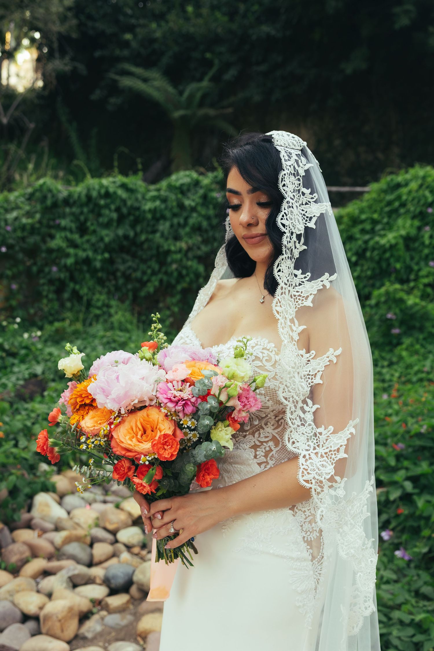Bride in lace veil and white gown holds colorful bouquet of orange and pink flowers in a garden setting.