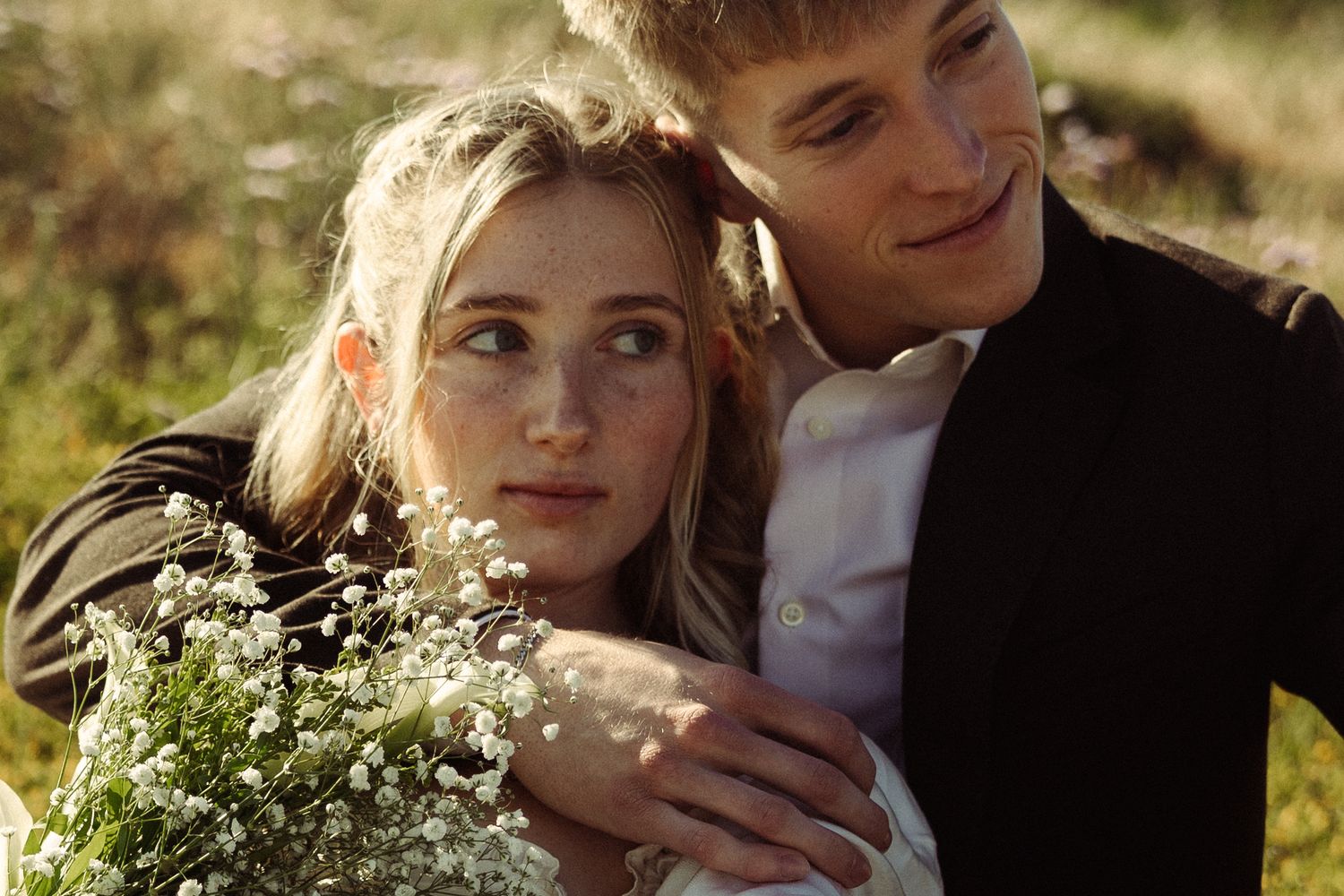 Romantic couple embraces in golden sunlight surrounded by lush greenery and flowers in an outdoor garden setting.