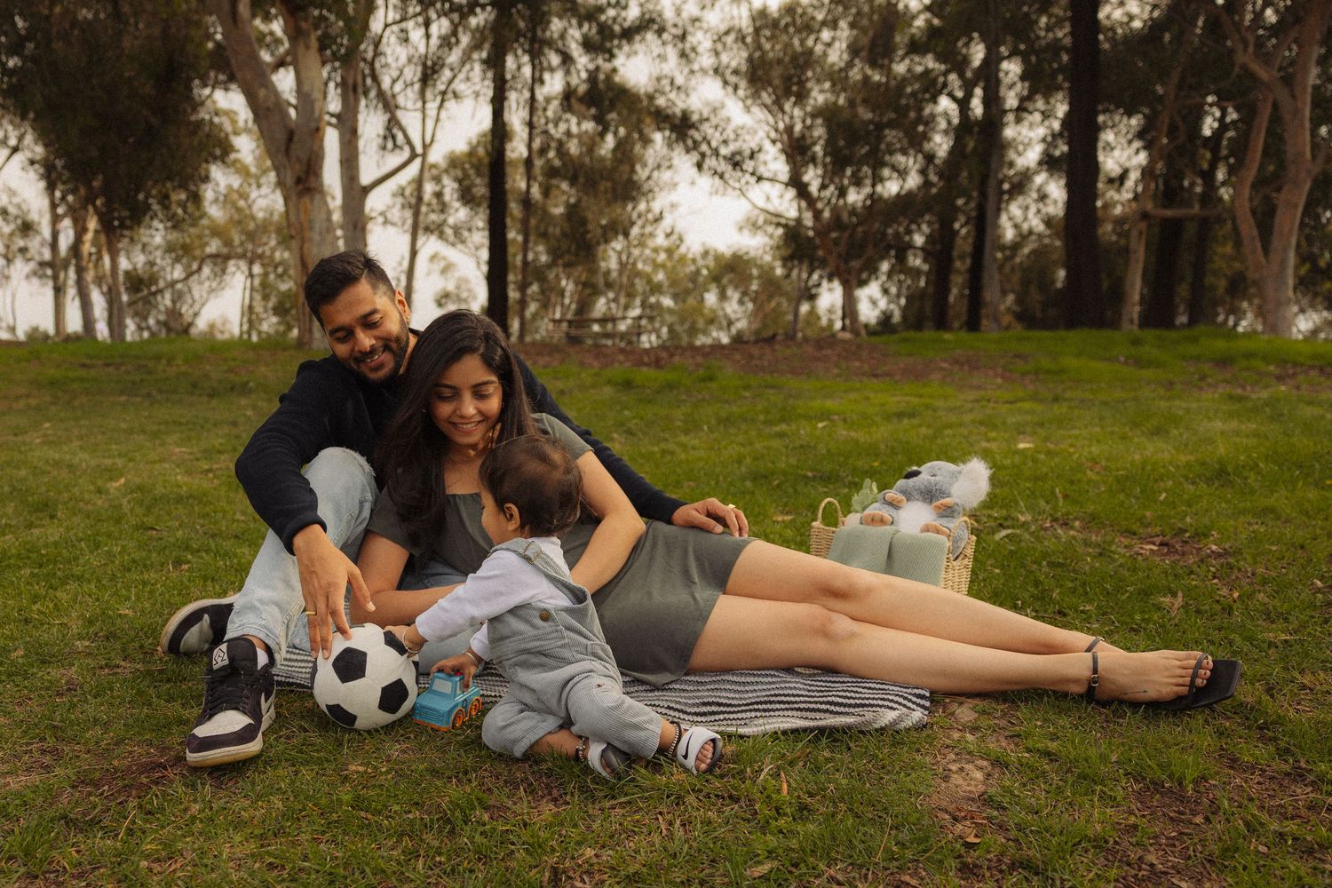 A couple relaxes on grass in a park with a soccer ball and picnic basket during a warm afternoon.
