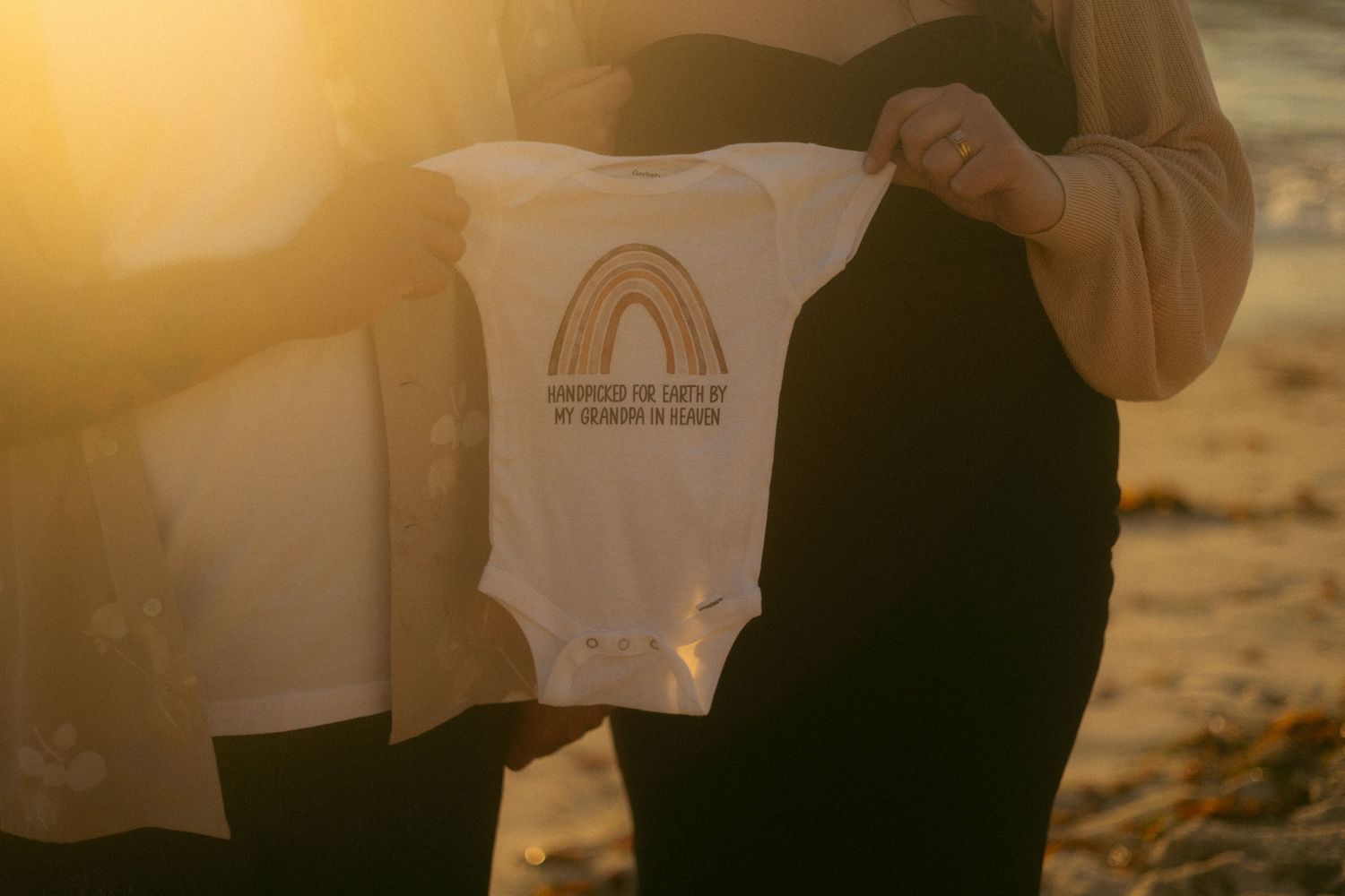 White baby onesie with rainbow design shown at golden sunset on beach.