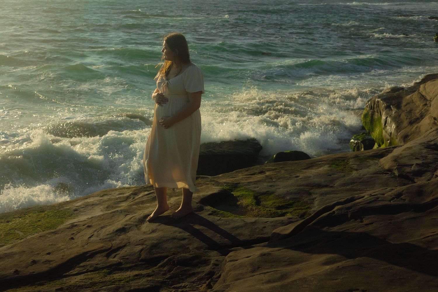 A person in a white dress stands on mossy coastal rocks as ocean waves crash behind them in warm evening light.