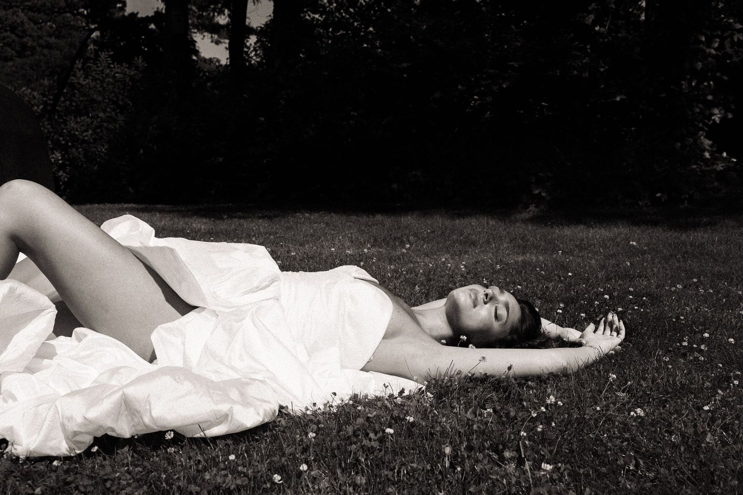 Black and white photo of a woman in a white dress lying on grass in a sunlit park.