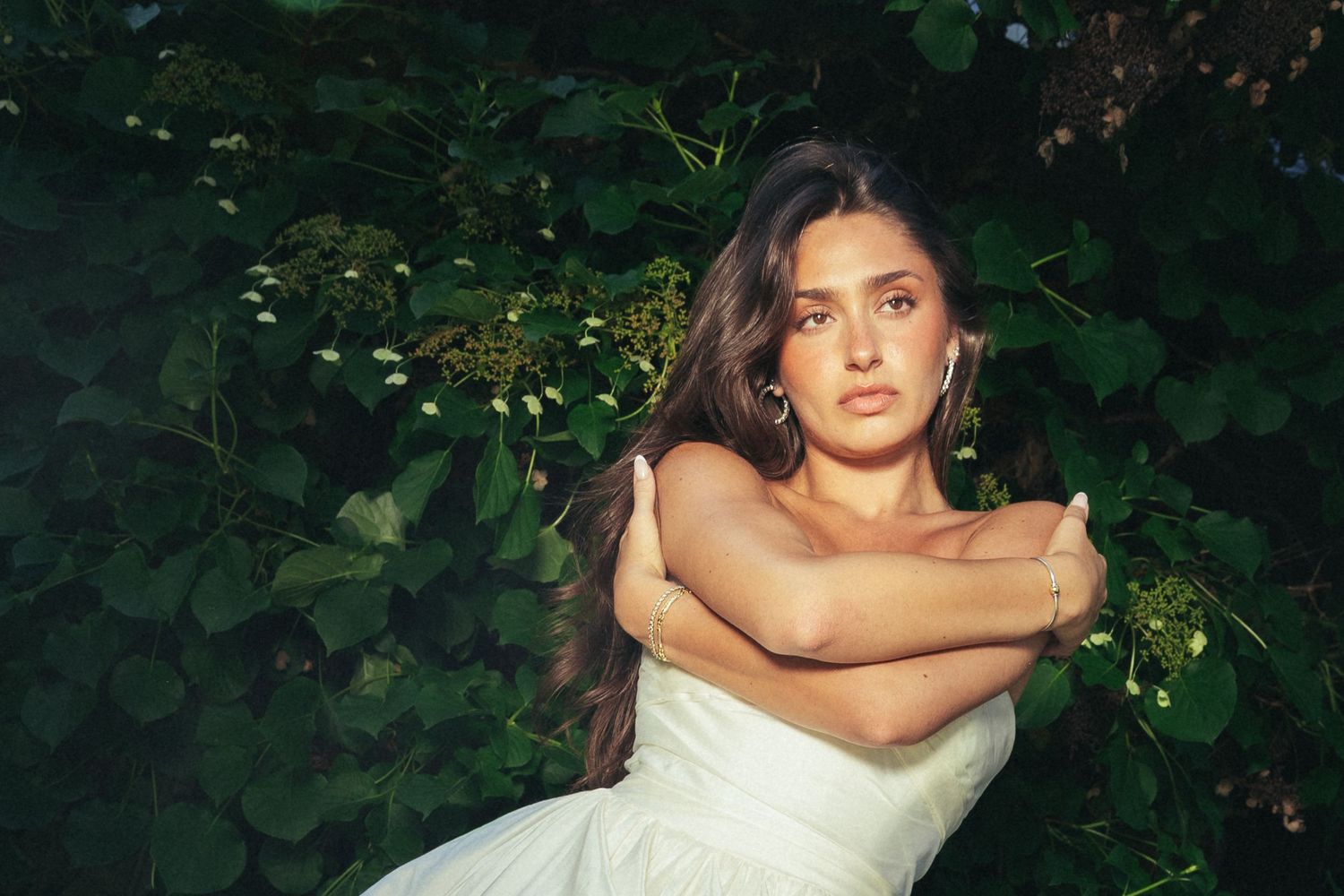 Young woman in white dress with crossed arms poses among lush green foliage outdoors.