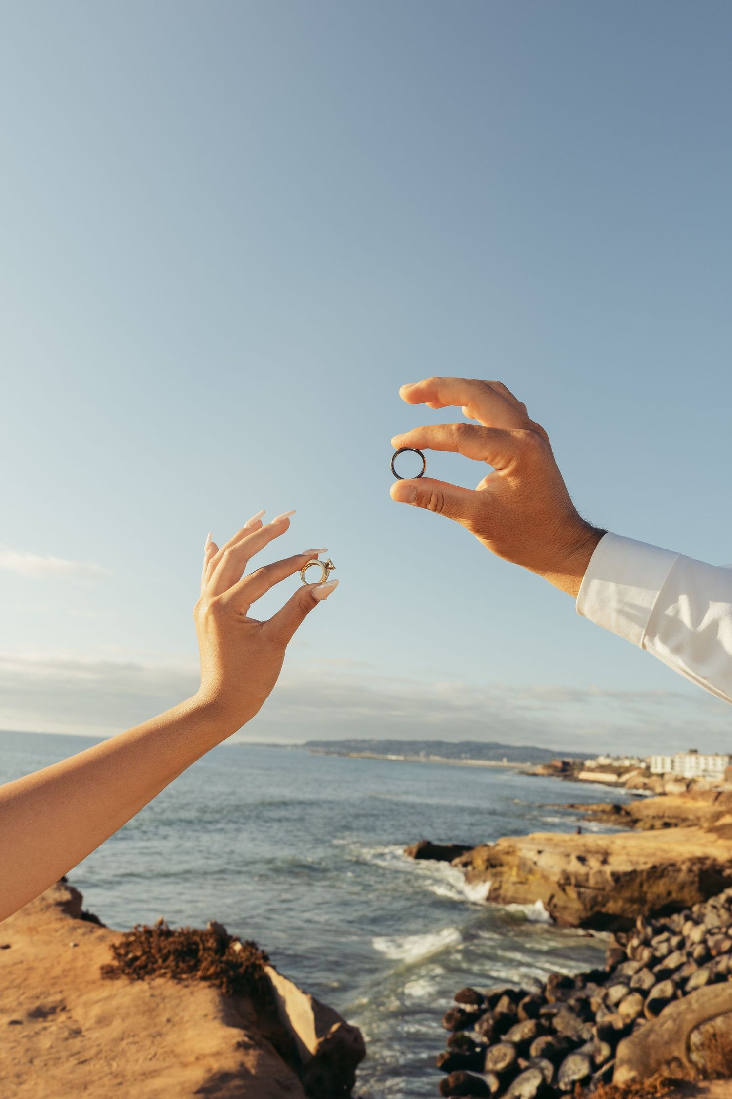 Two hands holding wedding rings near a rocky ocean coastline under a clear blue sky.