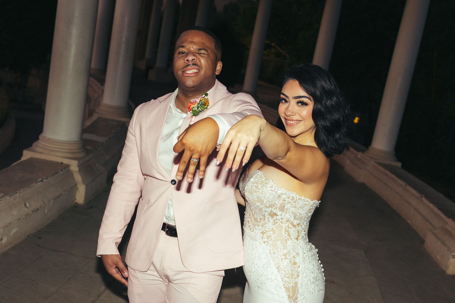 A couple in formal attire poses together, man in pink suit with colorful necklace, woman in beaded gown showing manicured nails.