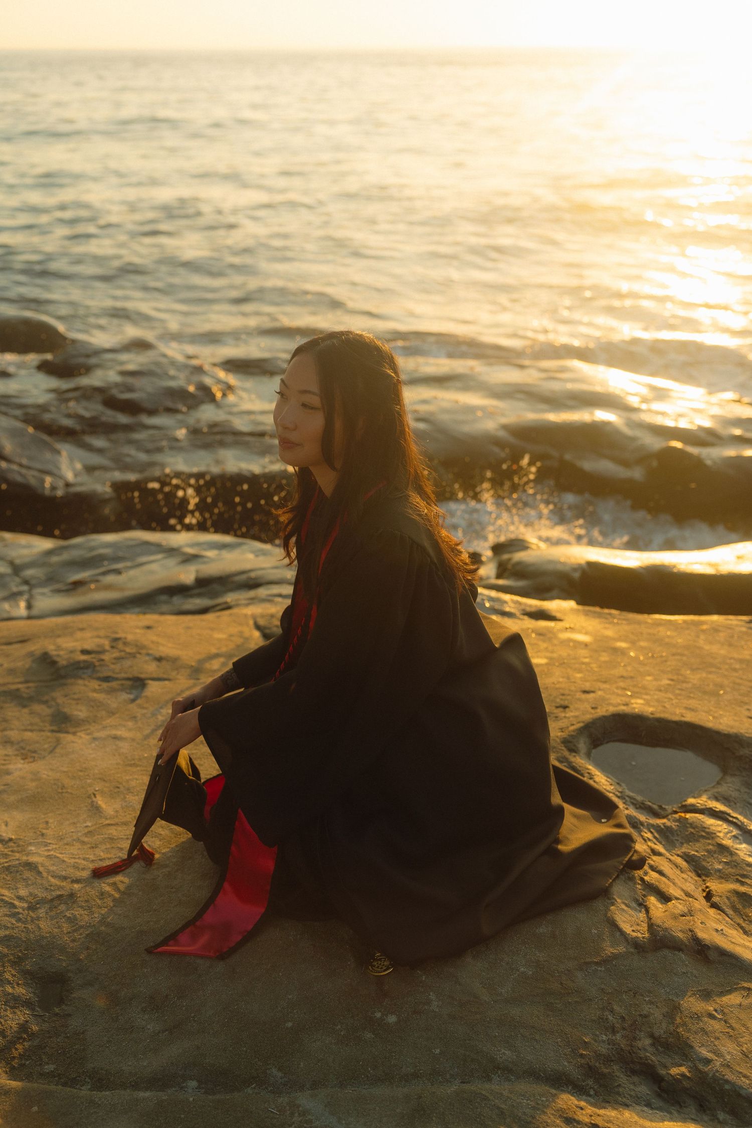 A silhouetted figure in black sits on coastal rocks at sunset while waves crash against the shore creating golden reflections.