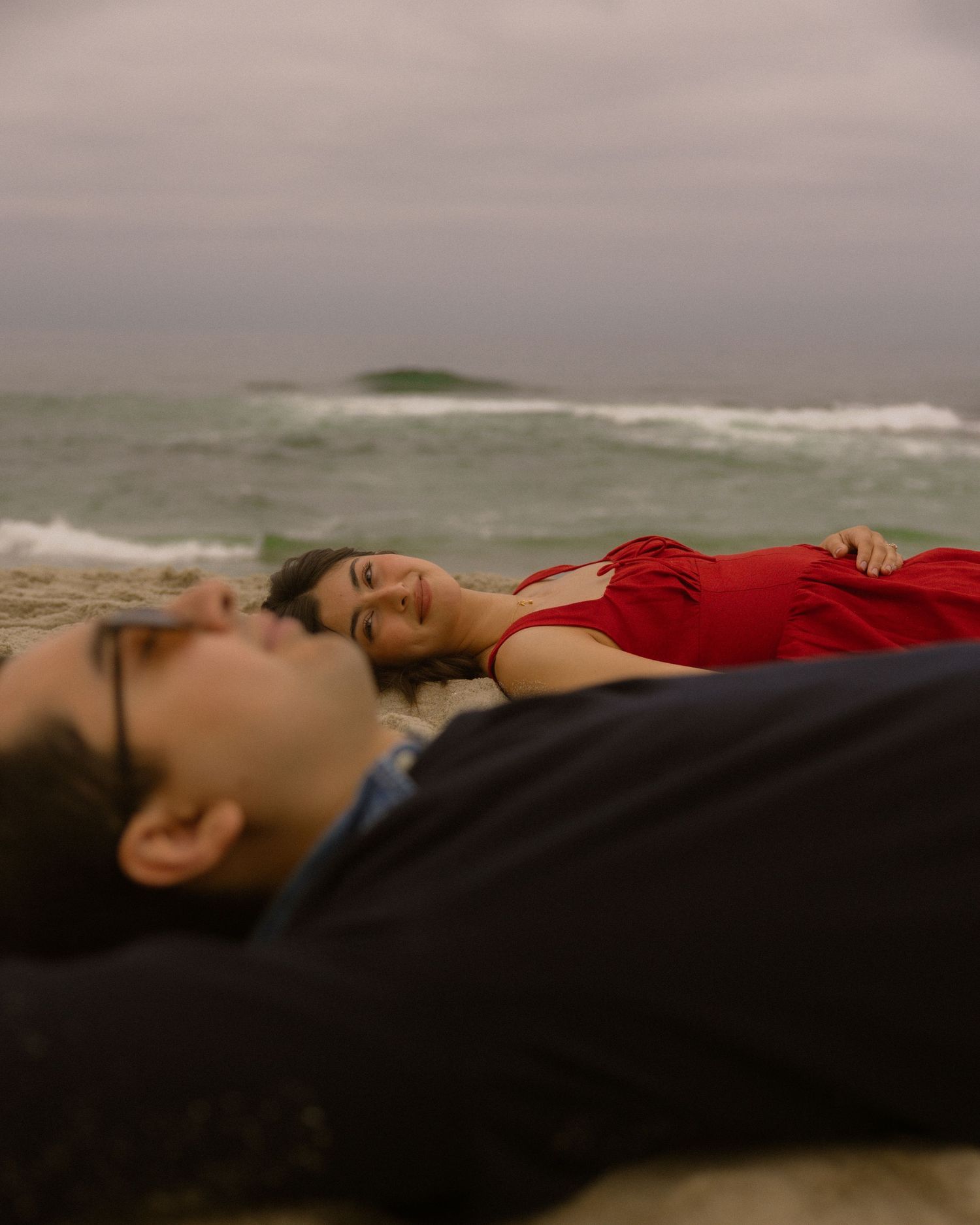 Person in red dress lying on dark surface against moody coastal backdrop at dusk.