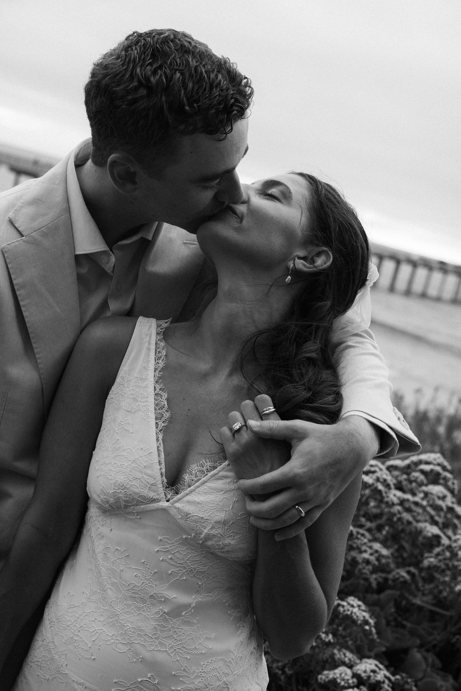 Black and white photo of a couple sharing a romantic kiss, bride in lace gown and groom in suit outdoors.