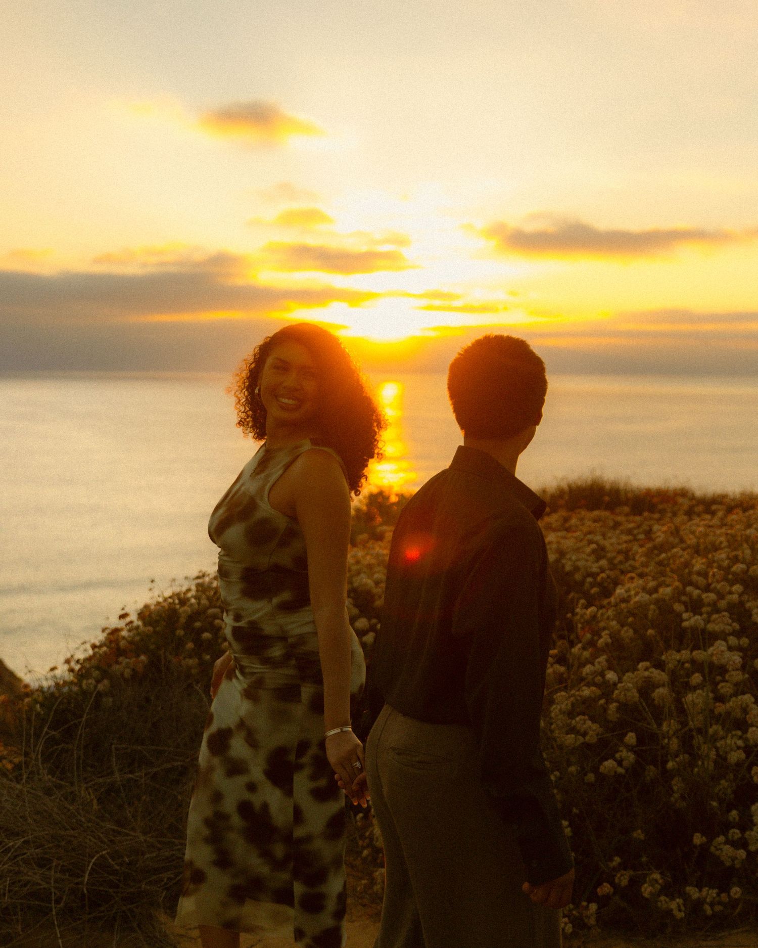 Silhouettes of people watching a golden sunset over the ocean with wildflowers in the foreground.