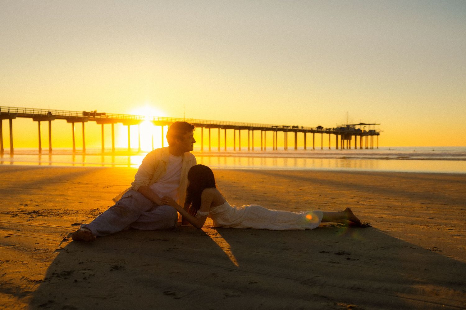 A silhouette of a person walking along a golden beach at sunset with a pier in the background.