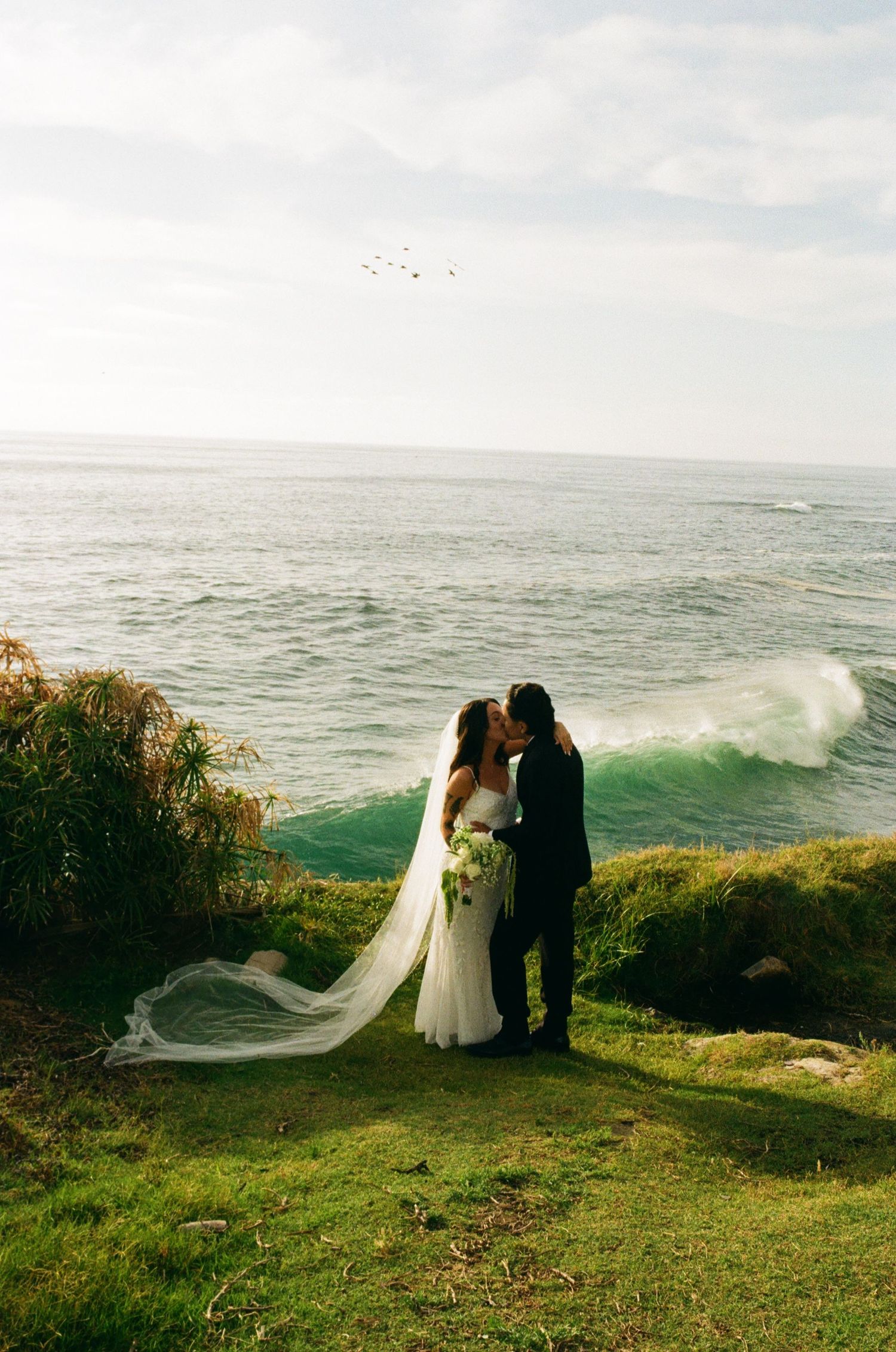 Bride and groom kissing on a coastal cliff with ocean waves and flowing wedding veil in the background.