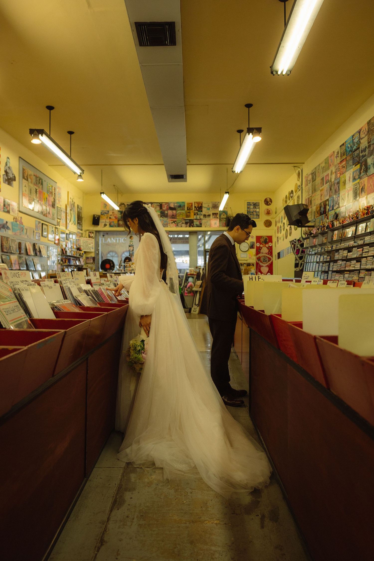 A bride in a flowing white wedding dress browses through a comic book store's shelves under fluorescent lighting.