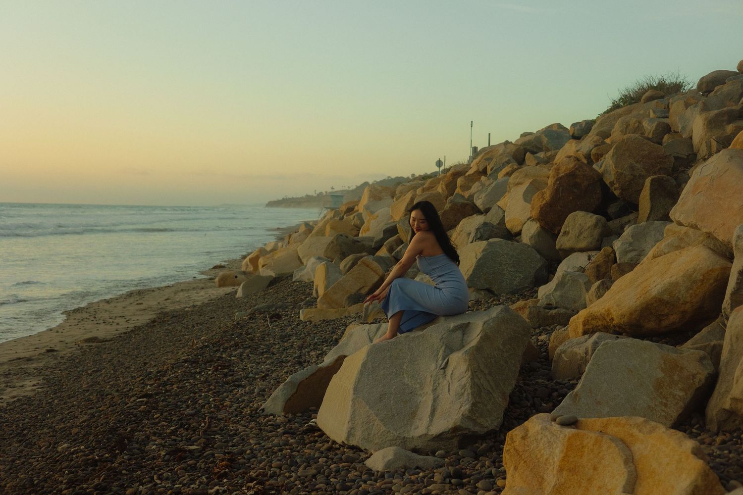 Someone sits along the rocky shore at sunset with large boulders and the ocean waves in view.