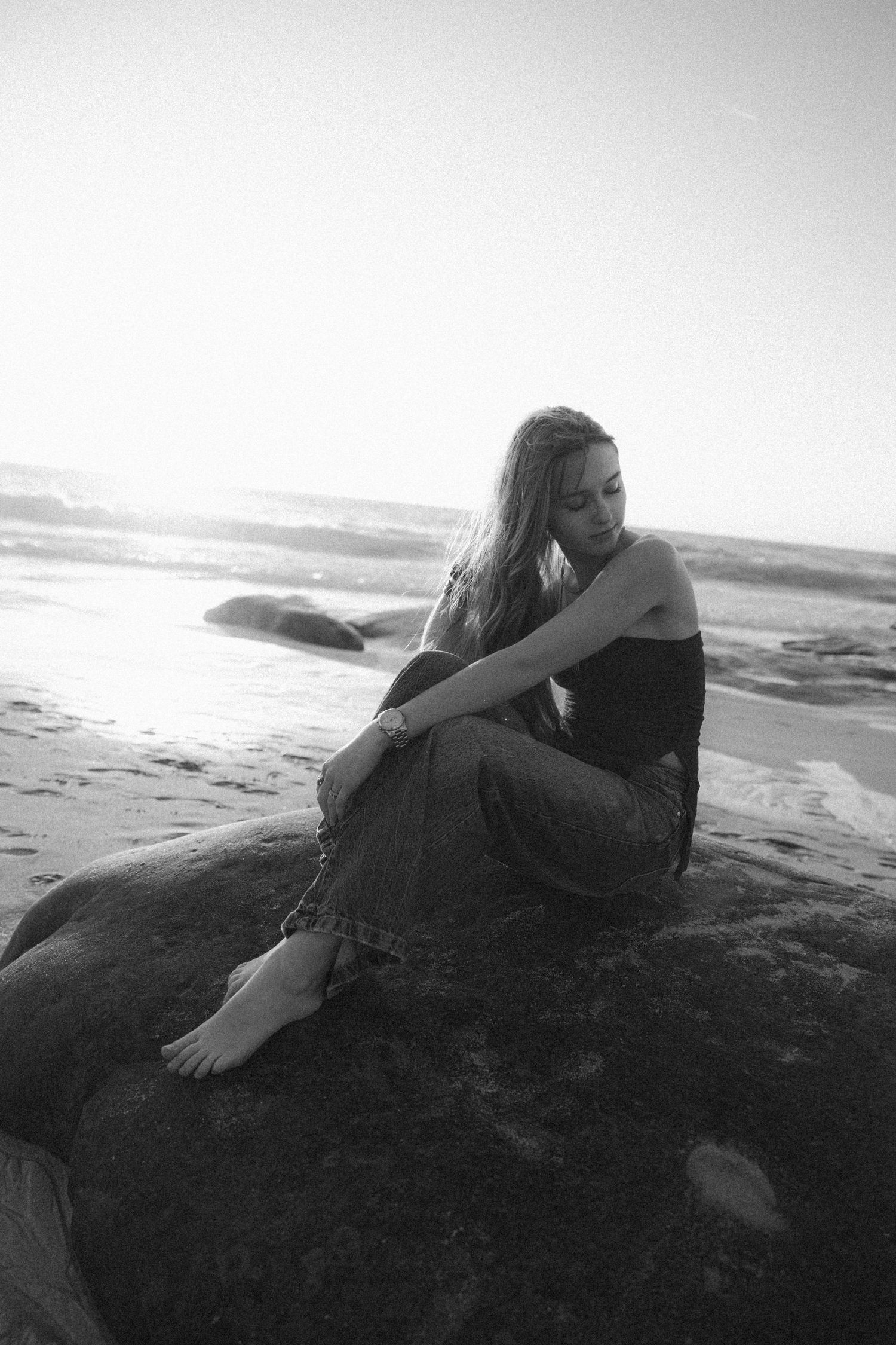 Black and white photo of a young woman sitting barefoot on a rock at the beach, gazing thoughtfully at the ocean.