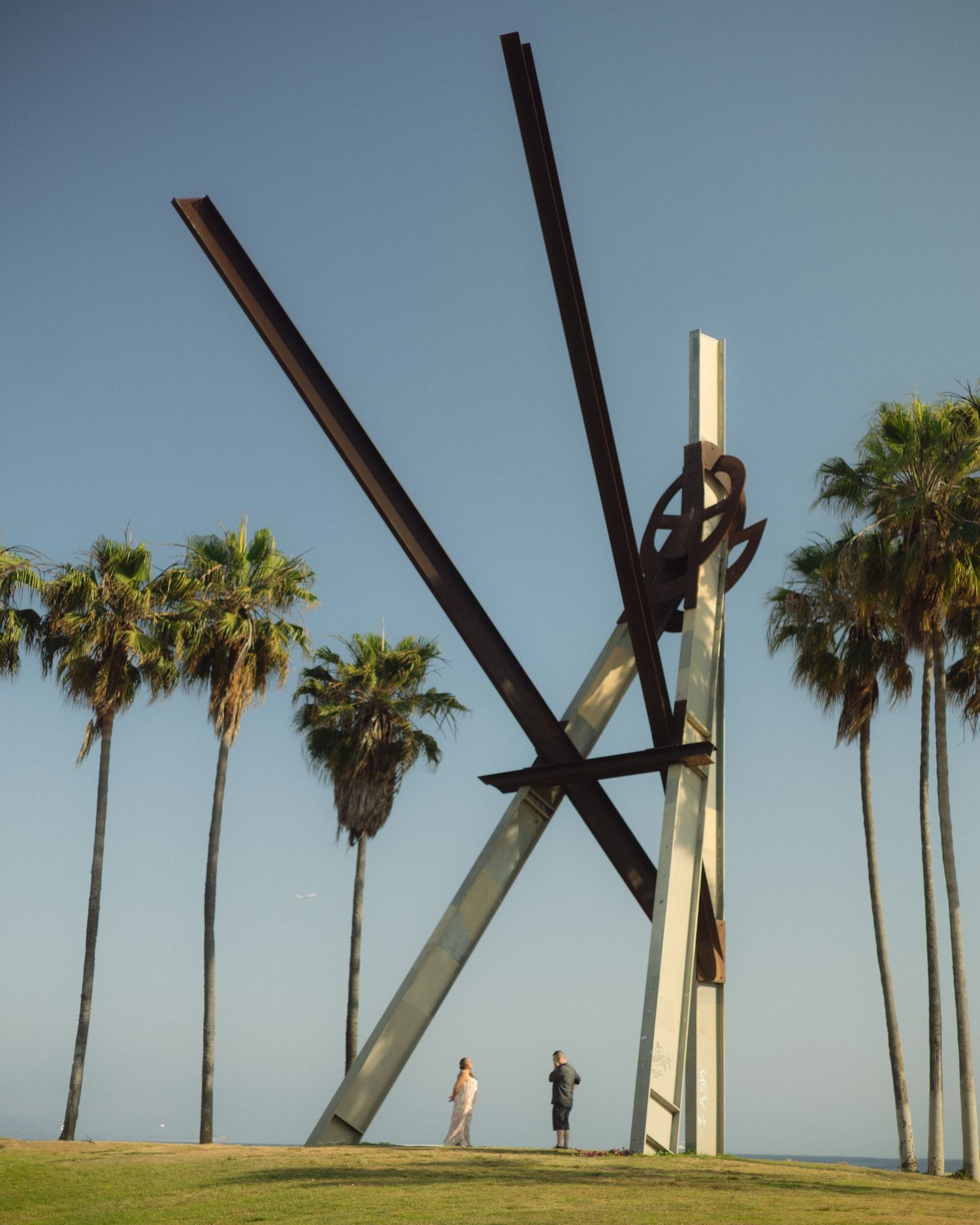 Large metal sculpture with crossing beams stands against blue sky with tall palm trees in Newport Beach, California.