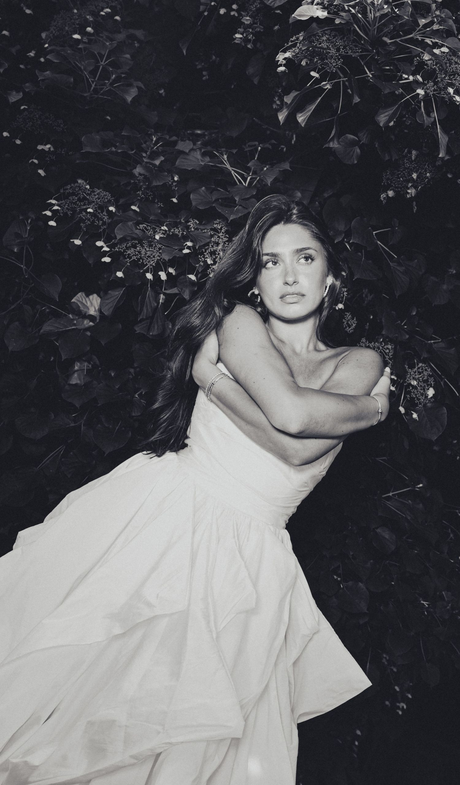 Black and white photo of a woman in a white strapless ball gown posing among lush foliage.
