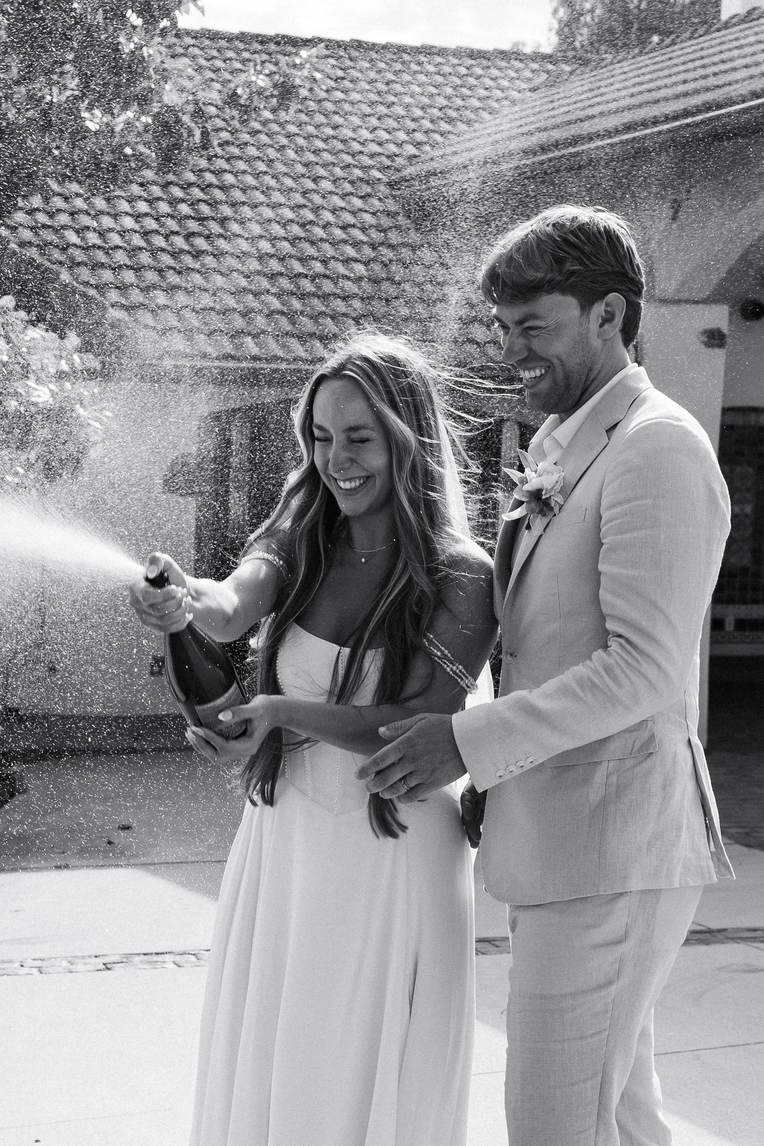Joyful newlyweds spraying champagne outdoors, bride in white dress and groom in beige suit, black and white photo.